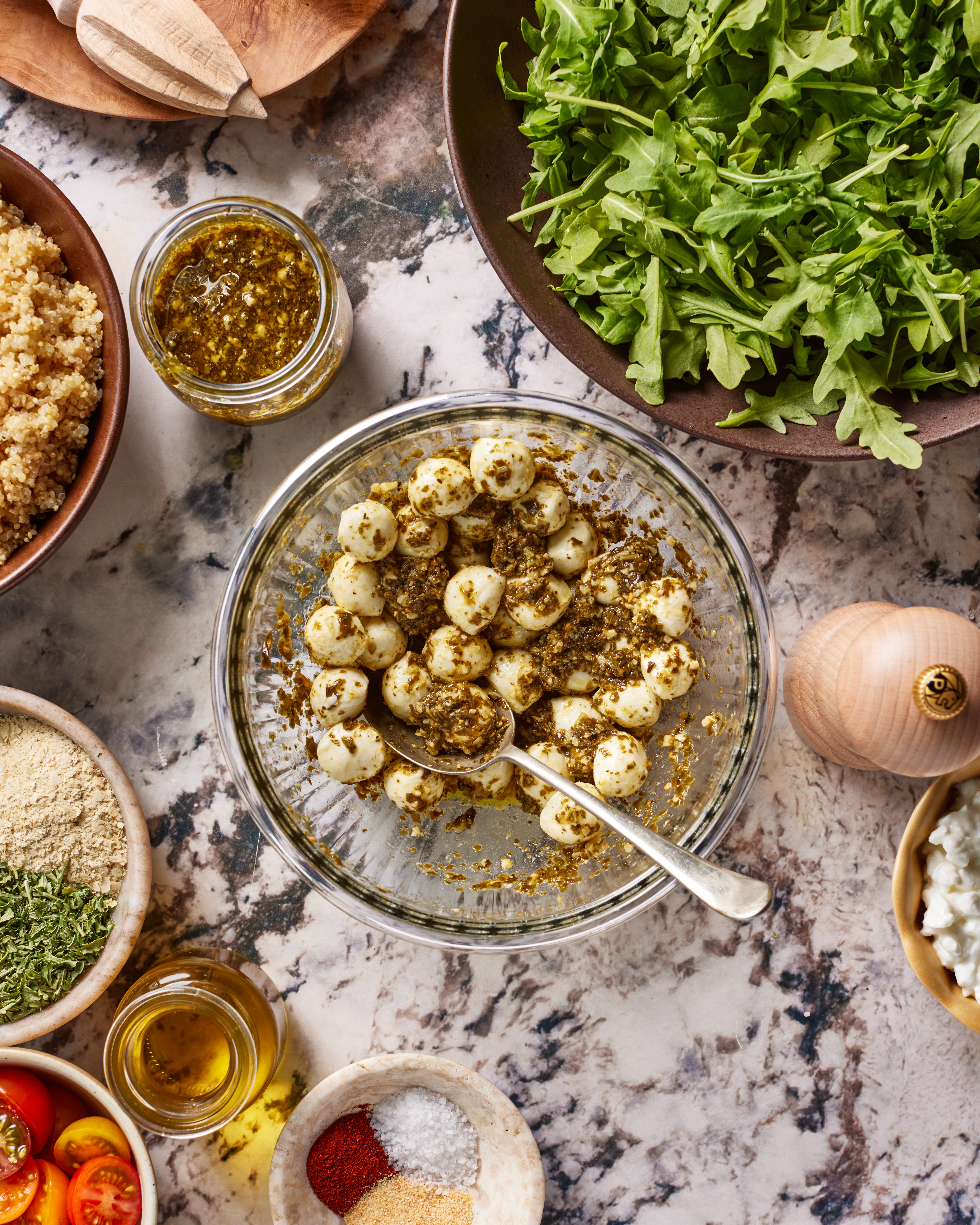 A glass bowl of marinated mozzarella balls with a spoon, surrounded by bowls of arugula, quinoa, olive oil, cherry tomatoes, spices, and grated cheese on a marble countertop.