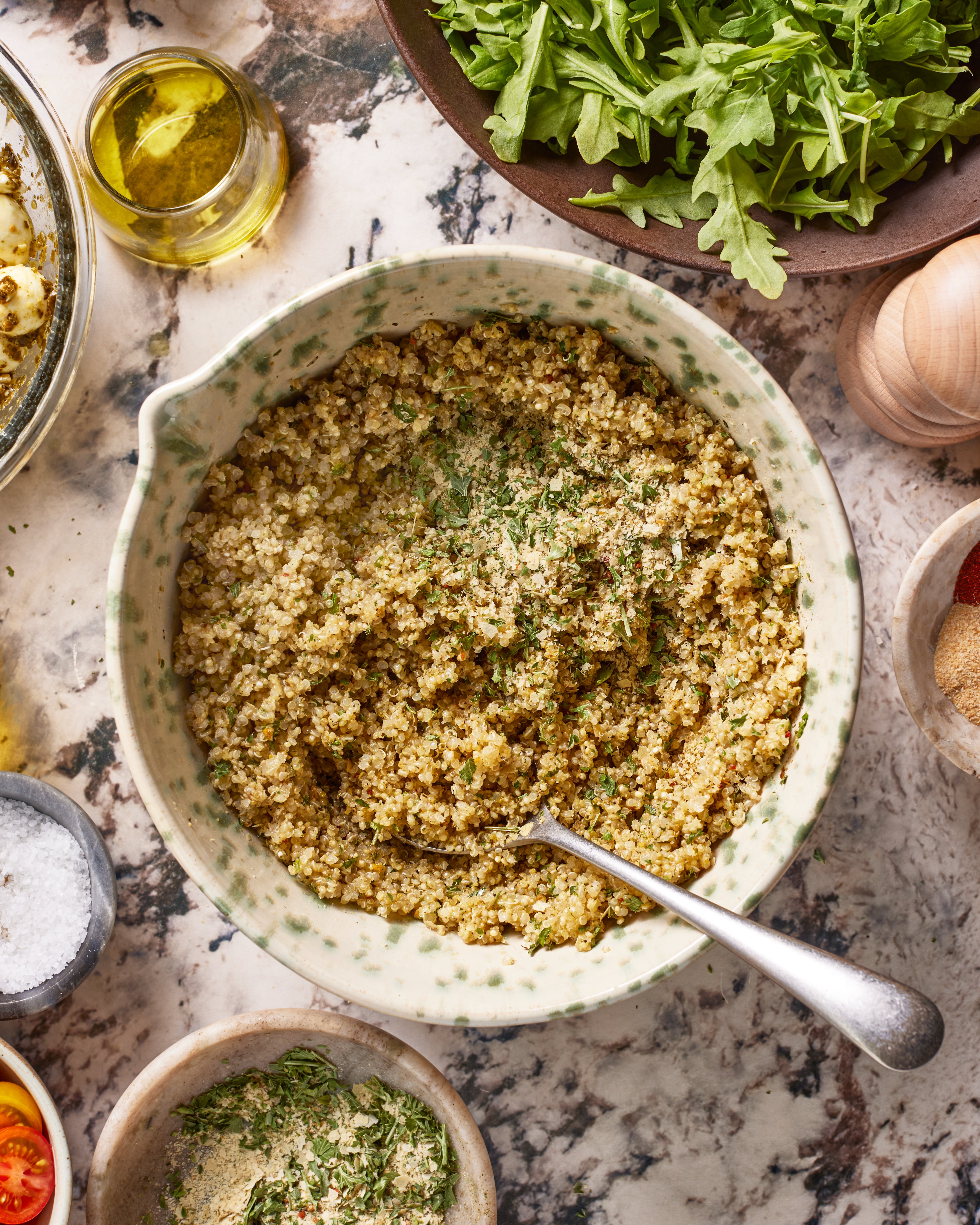 A bowl of cooked quinoa garnished with herbs sits on a marble surface, surrounded by fresh arugula, olive oil, a pepper grinder, tomatoes, and small bowls of seasonings. A spoon rests in the quinoa.