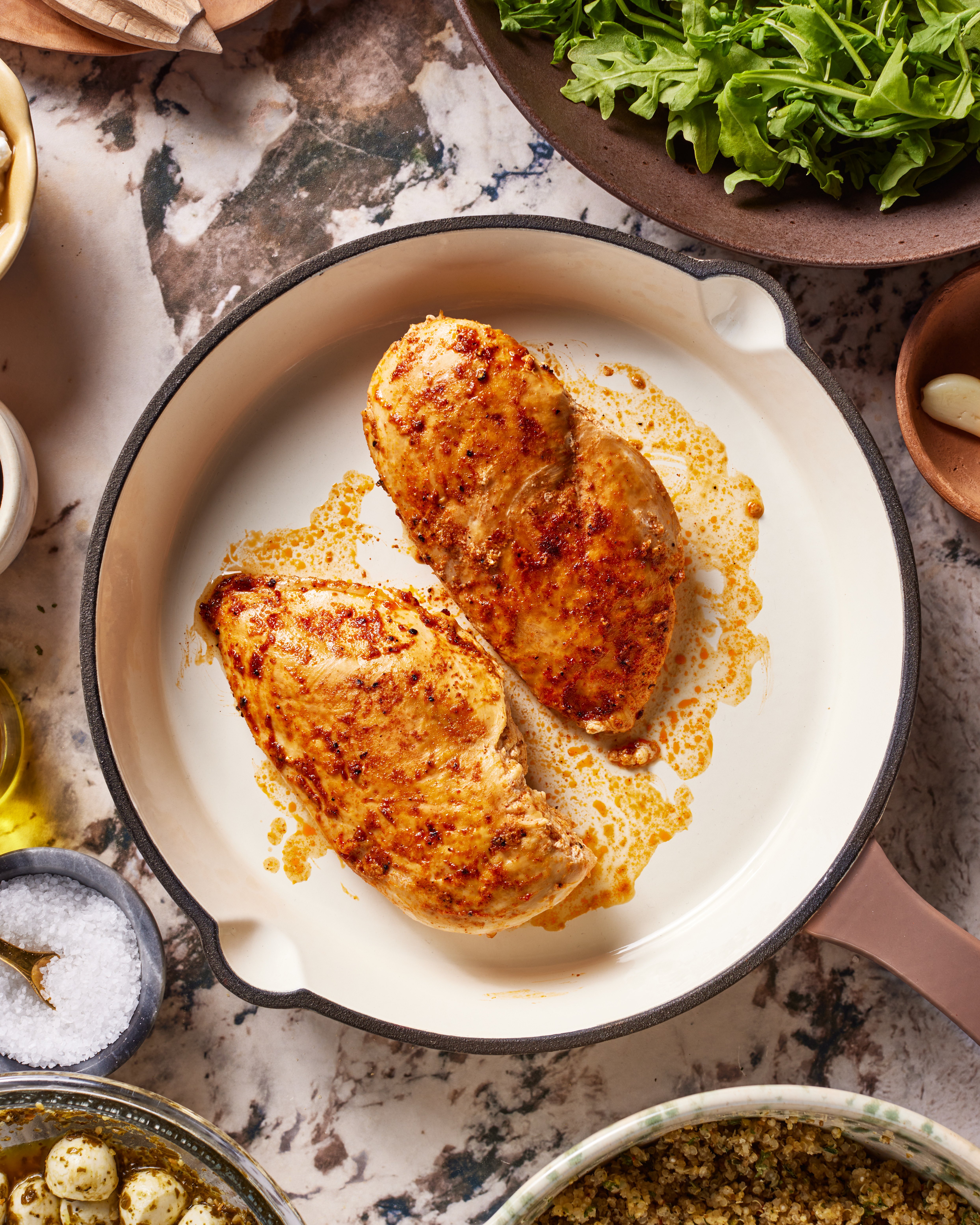 Two seasoned, cooked chicken breasts in a white skillet, surrounded by bowls of fresh greens, salt, oil, garlic, and other ingredients on a marble countertop.