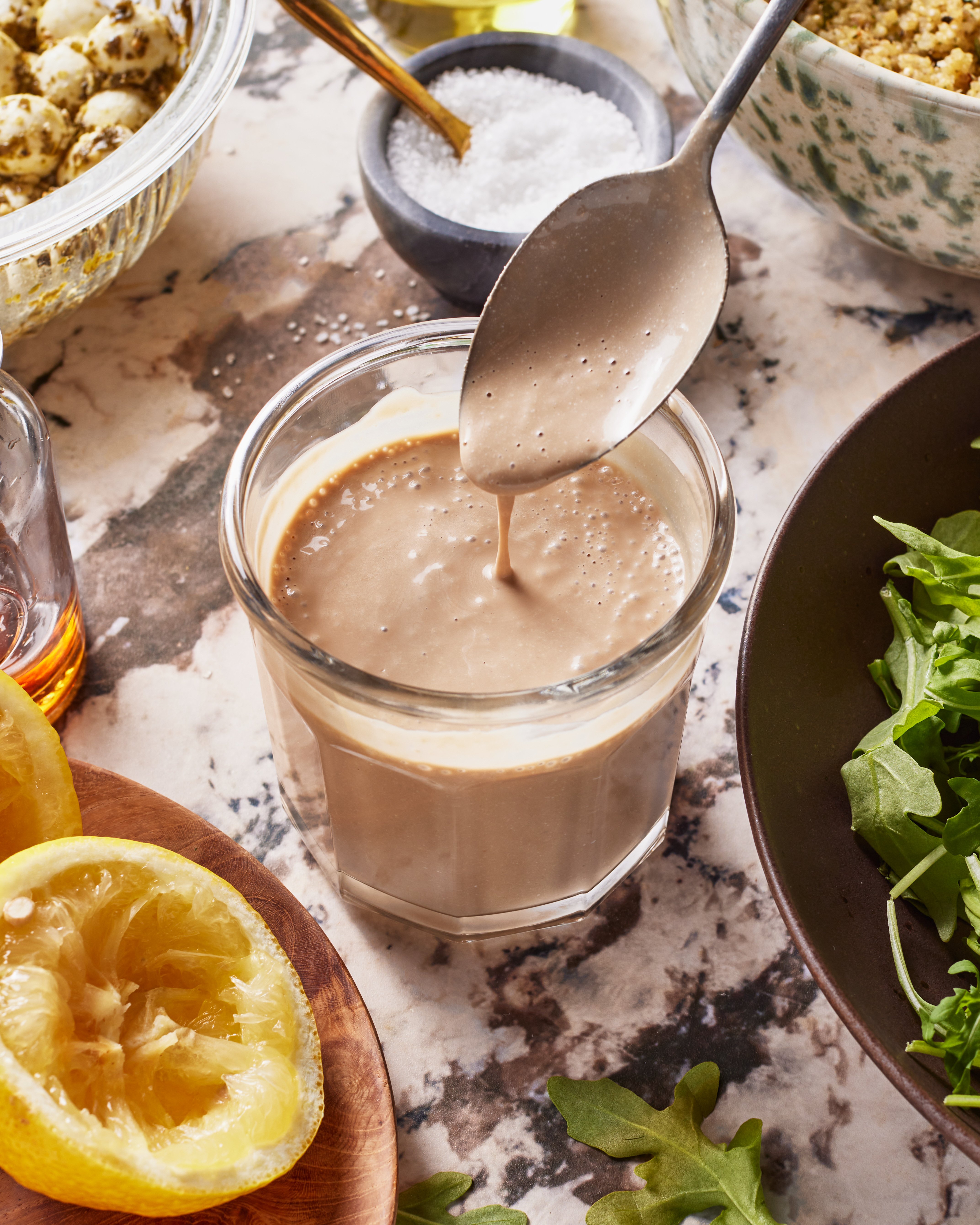 A spoon drizzles creamy dressing into a glass, surrounded by halved lemons, a bowl of arugula, a bowl of grains, and a small dish of salt on a marbled countertop. (Copycat Sweetgreen Chicken Caprese Salad)