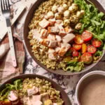 A Copycat Sweetgreen Chicken Caprese Bowl filled with quinoa, grilled chicken, cherry tomatoes, arugula, mozzarella balls, and creamy dressing sit on a marble surface next to a napkin, forks, and a bowl of extra dressing.