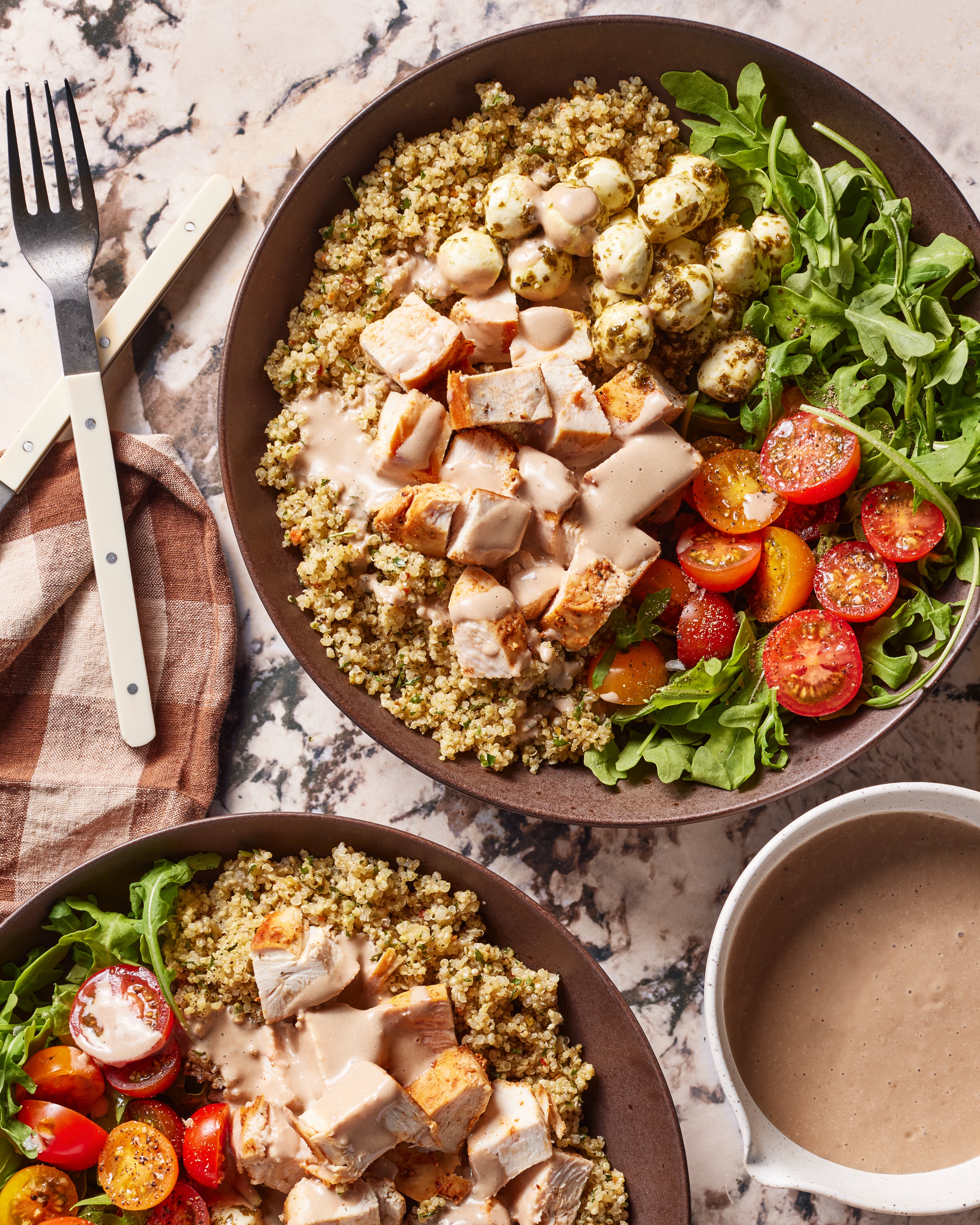 A Copycat Sweetgreen Chicken Caprese Bowl filled with quinoa, grilled chicken, cherry tomatoes, arugula, mozzarella balls, and creamy dressing sit on a marble surface next to a napkin, forks, and a bowl of extra dressing.