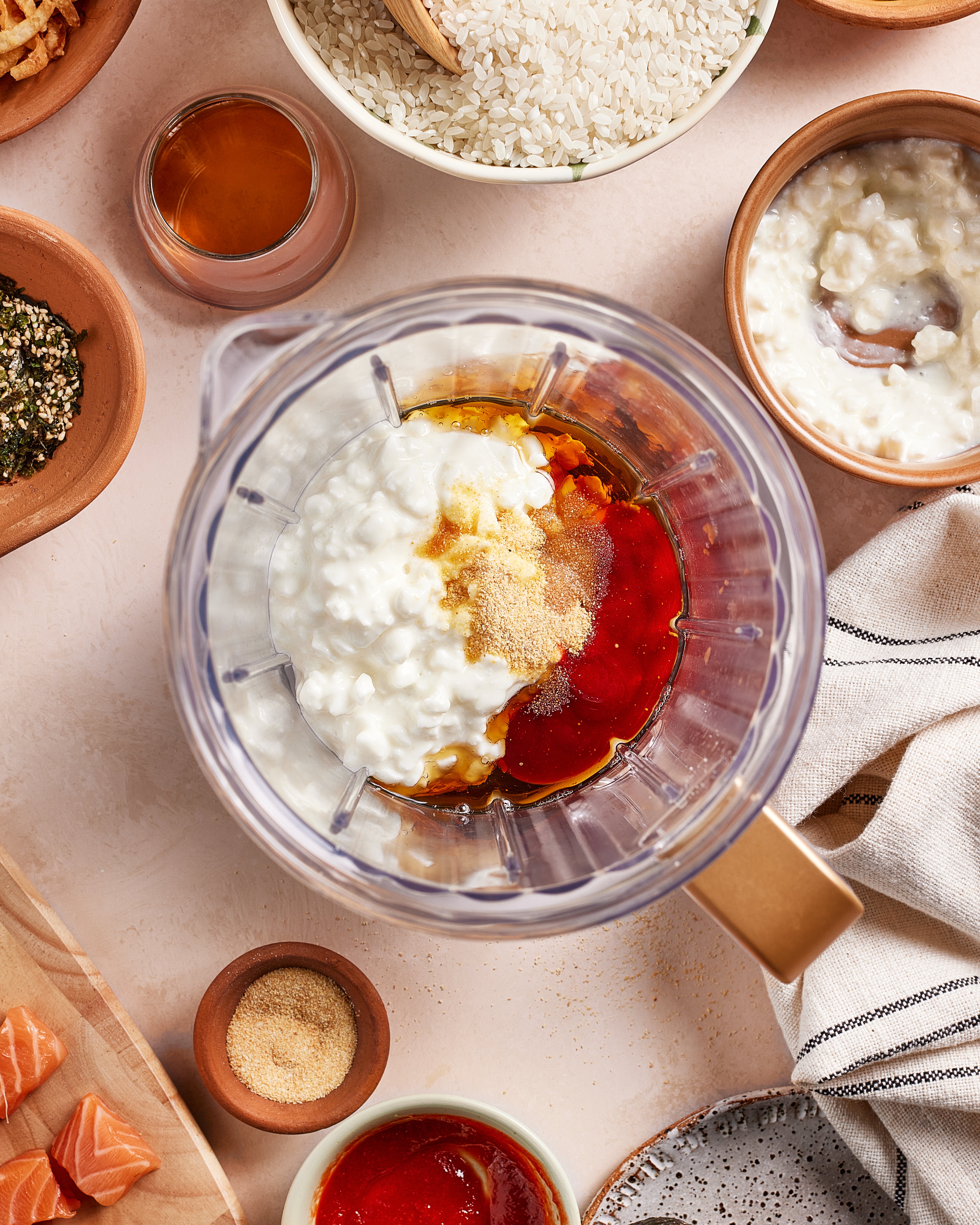A blender filled with cottage cheese, hot sauce, and spices sits on a table surrounded by bowls of rice, seasonings, sauce, and cubes of raw salmon, ready for meal preparation.