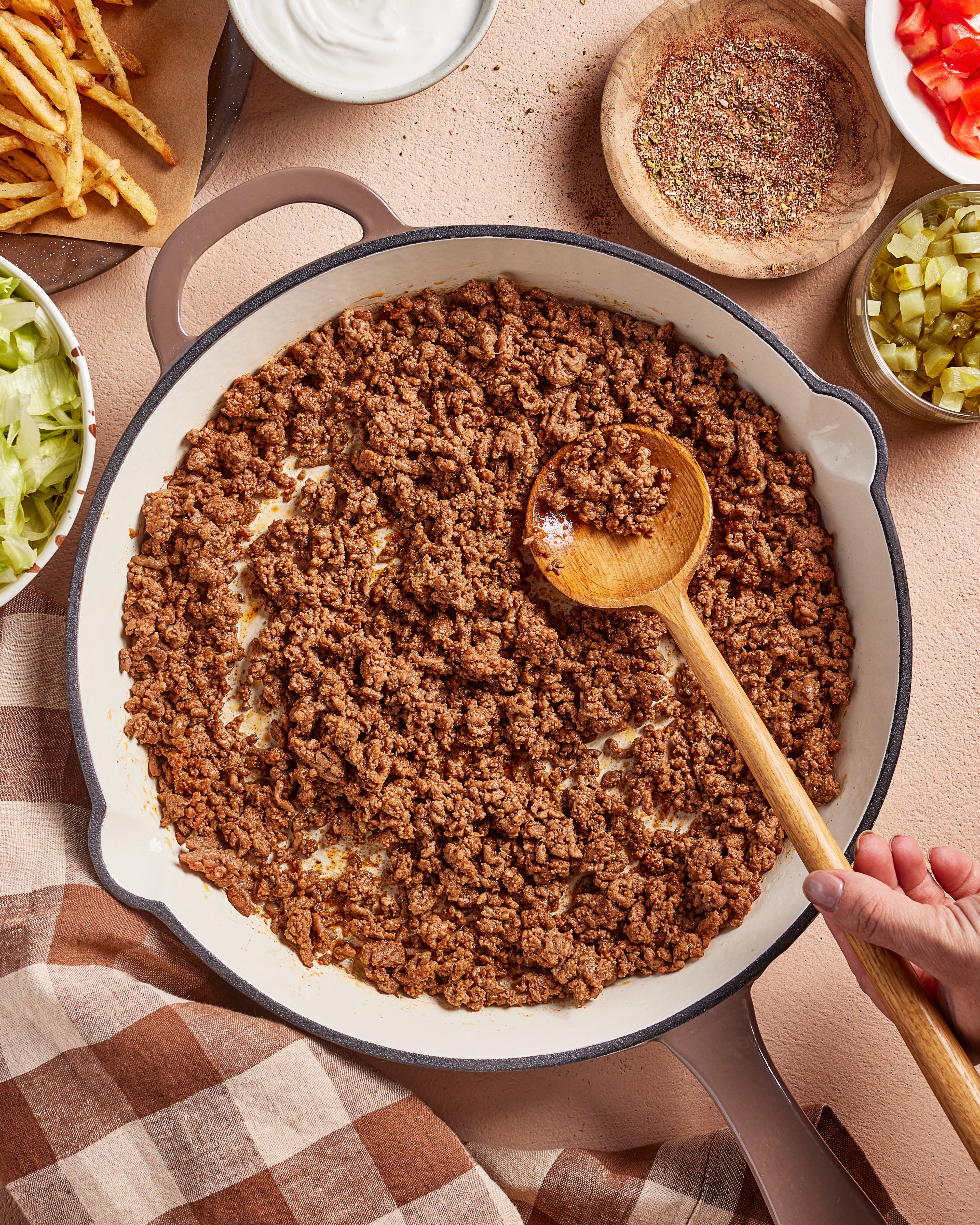 A skillet filled with cooked ground beef being stirred with a wooden spoon, surrounded by bowls of shredded lettuce, pickles, French fries, shredded cheese, sour cream, and spices on a beige surface with a checkered cloth.