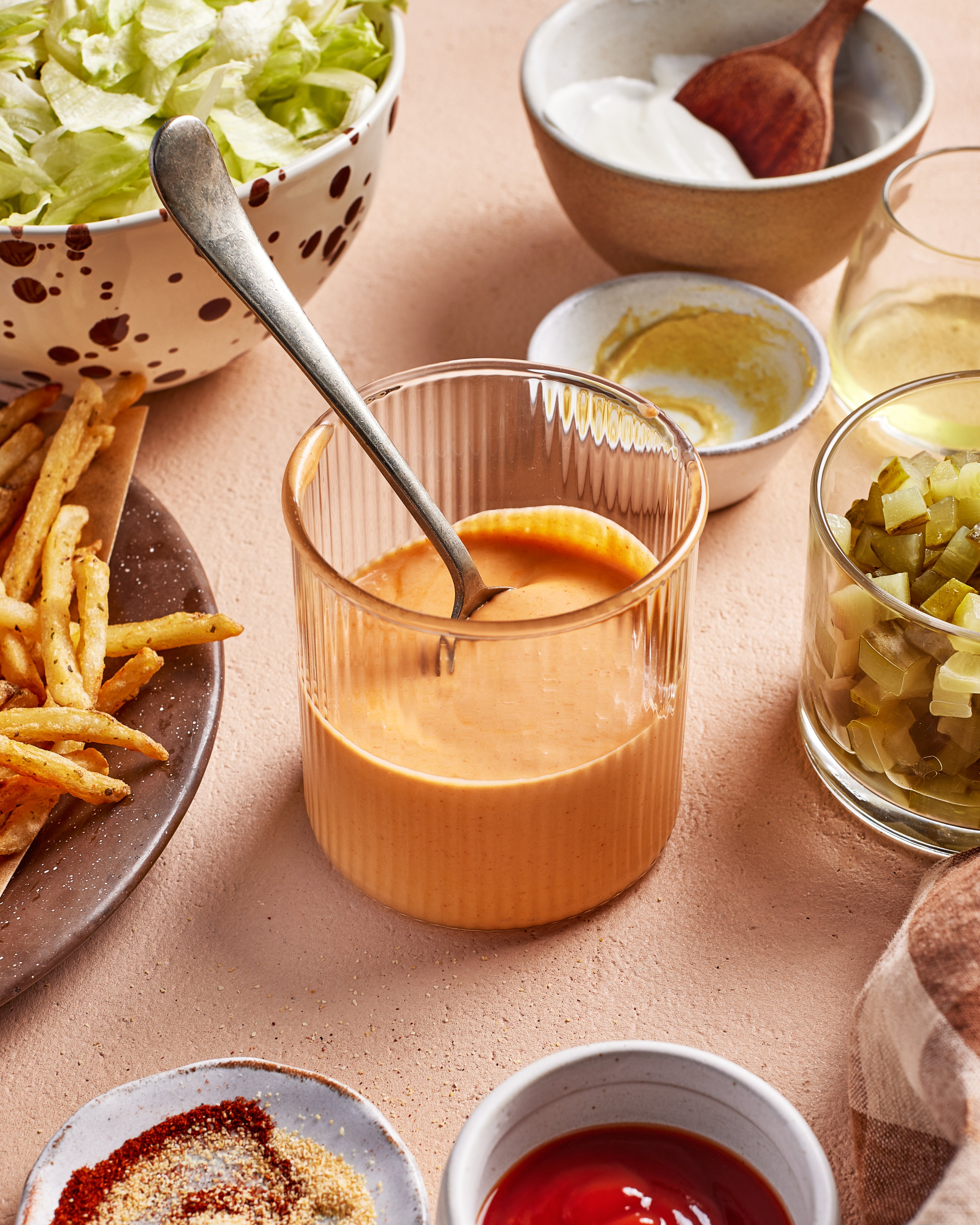 A glass with orange-colored sauce and a spoon sits on a table surrounded by bowls of lettuce, pickles, spices, ketchup, fries, and a bowl with a wooden spoon, all arranged on a light surface.