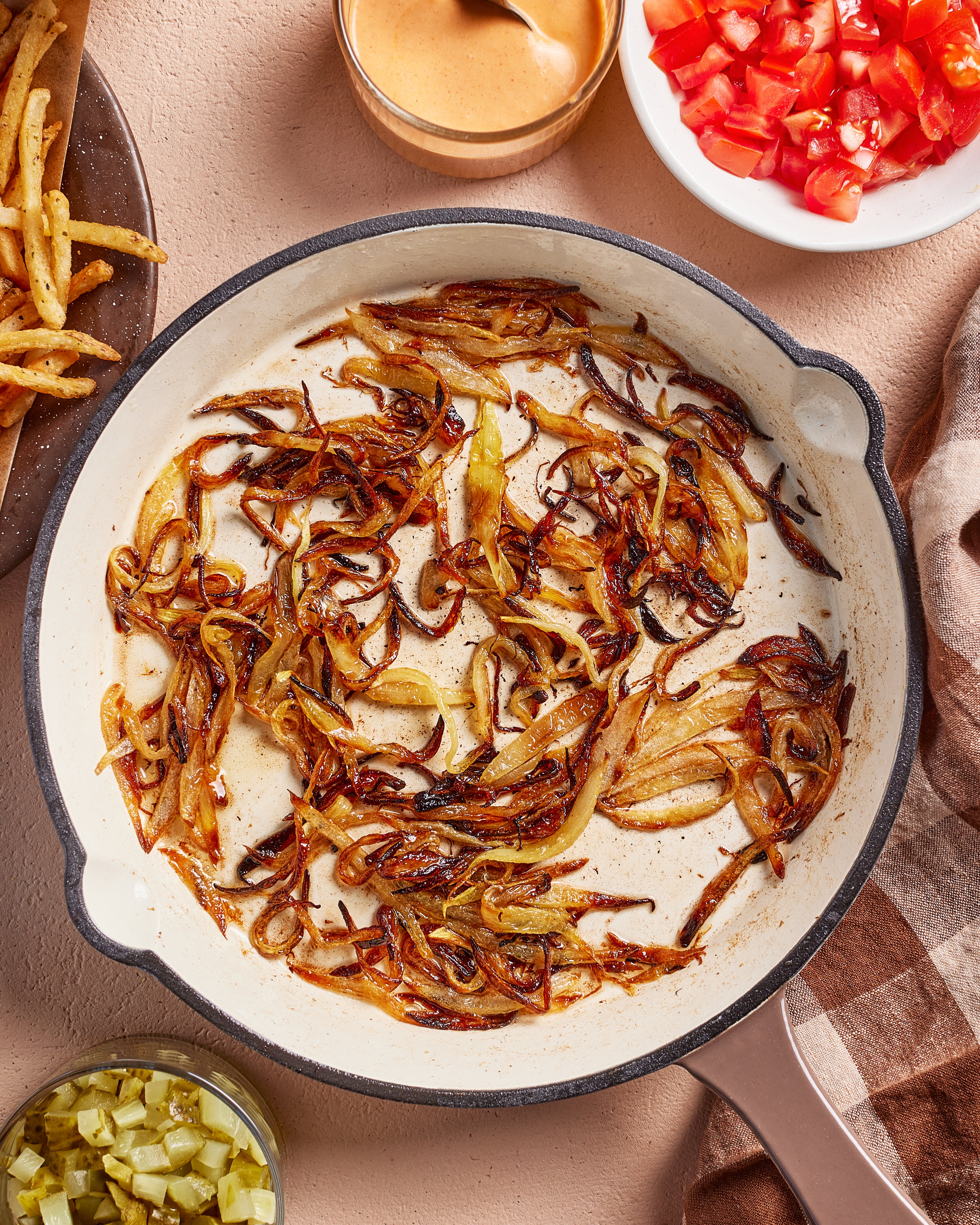 A skillet with caramelized onions sits on a table, surrounded by bowls of diced tomatoes, pickles, sauce, French fries, and a checkered towel.