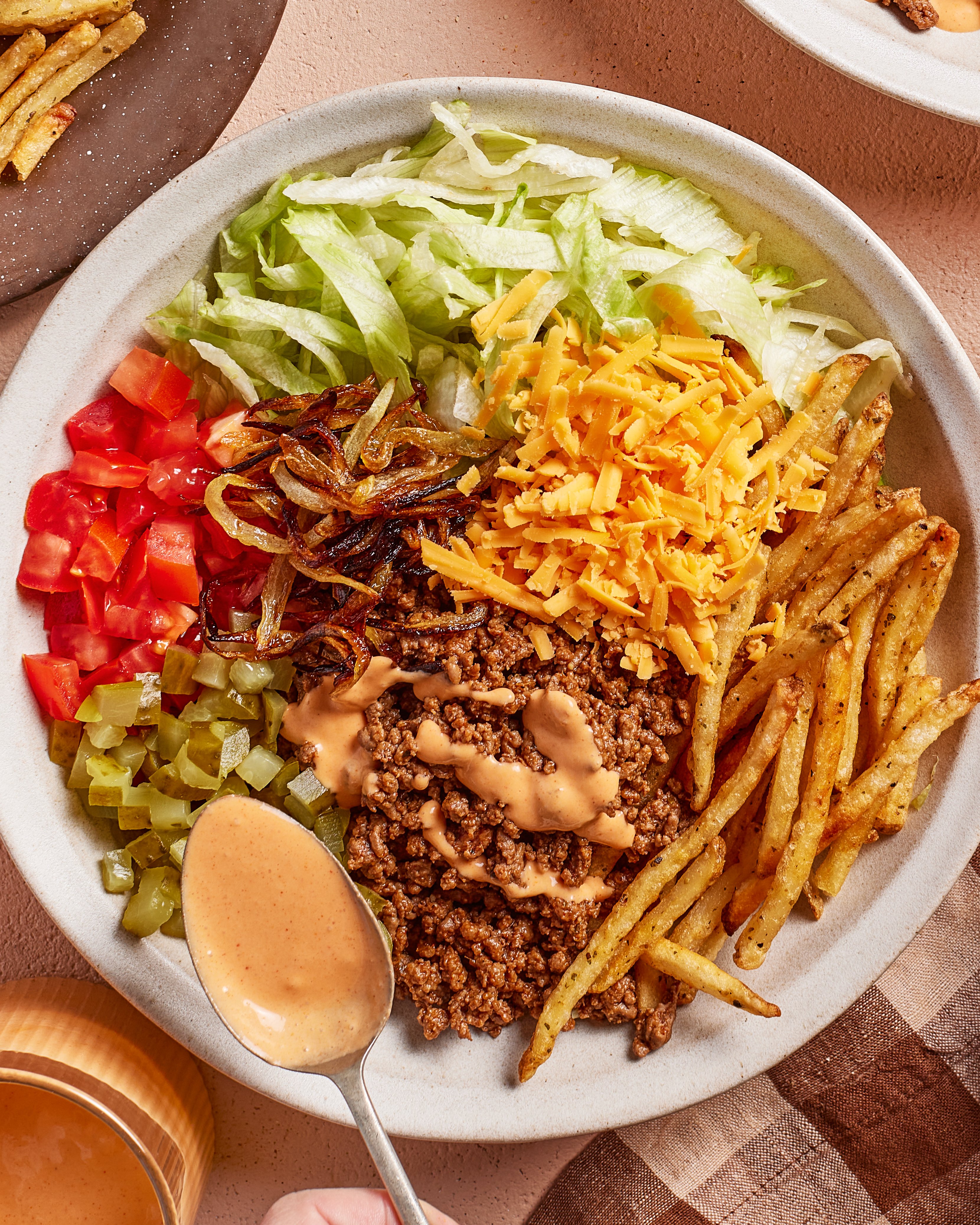 A bowl filled with shredded lettuce, diced tomatoes, pickles, caramelized onions, shredded cheddar cheese, seasoned ground beef, crinkle-cut fries, and sauce being drizzled on top with a spoon.