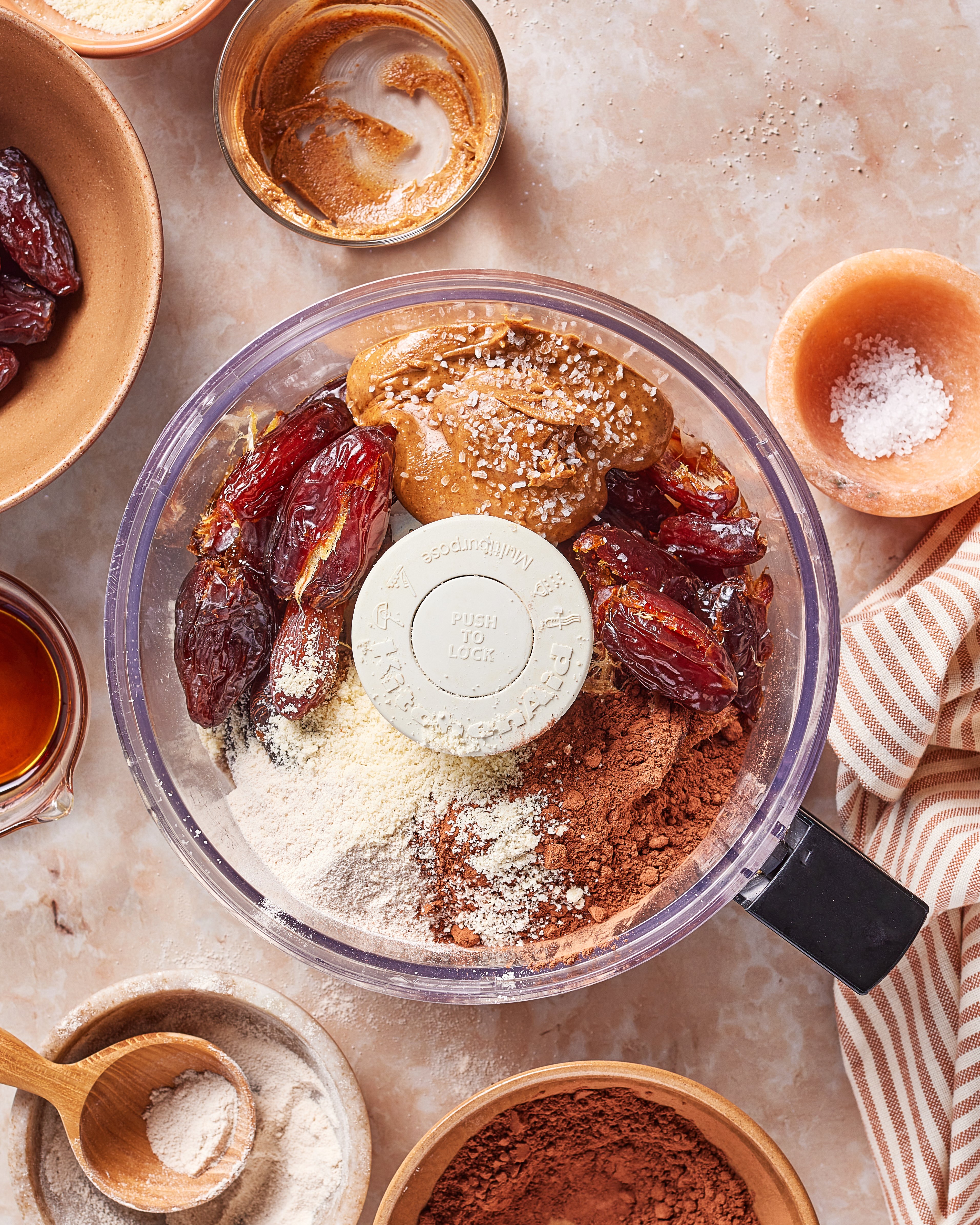 A food processor bowl filled with dates, peanut butter, cocoa powder, almond flour, and a sprinkle of salt, surrounded by small bowls of ingredients on a marble surface.