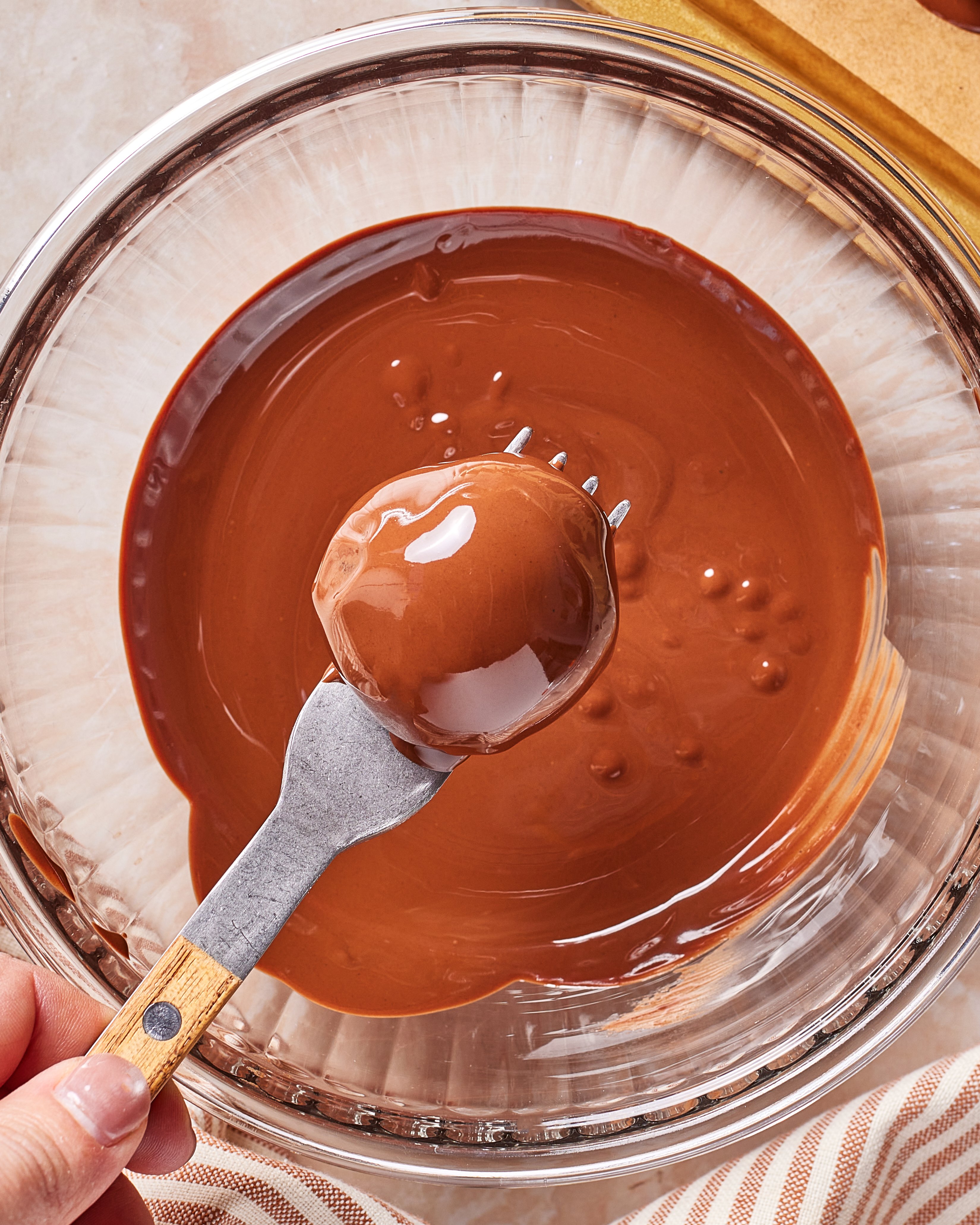 A hand holds a fork with a round treat coated in melted chocolate over a glass bowl filled with more melted chocolate.