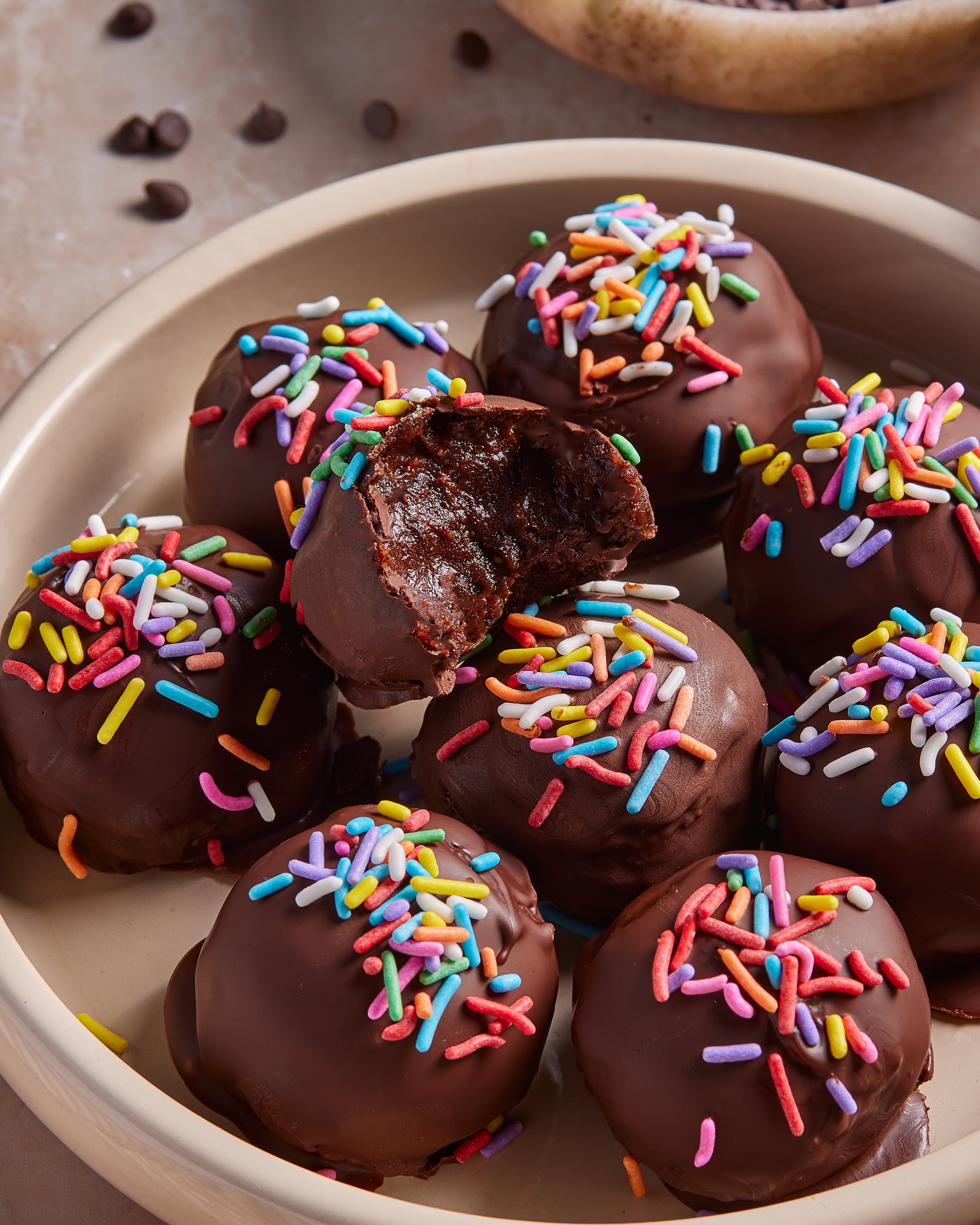 A plate of chocolate-coated dessert balls topped with colorful rainbow sprinkles. One ball has a bite taken out, showing a rich, fudgy interior. The plate sits on a light-colored surface with some scattered chocolate chips.