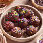 A bowl filled with chocolate-coated balls topped with colorful sprinkles sits on a marble surface, surrounded by bowls of chocolate chips, cocoa powder, and a striped cloth. (Viral High Protein Brownie Bites)