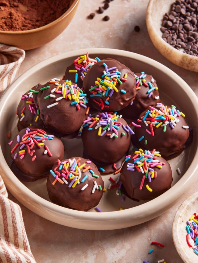 A bowl filled with chocolate-coated balls topped with colorful sprinkles sits on a marble surface, surrounded by bowls of chocolate chips, cocoa powder, and a striped cloth. (Viral High Protein Brownie Bites)