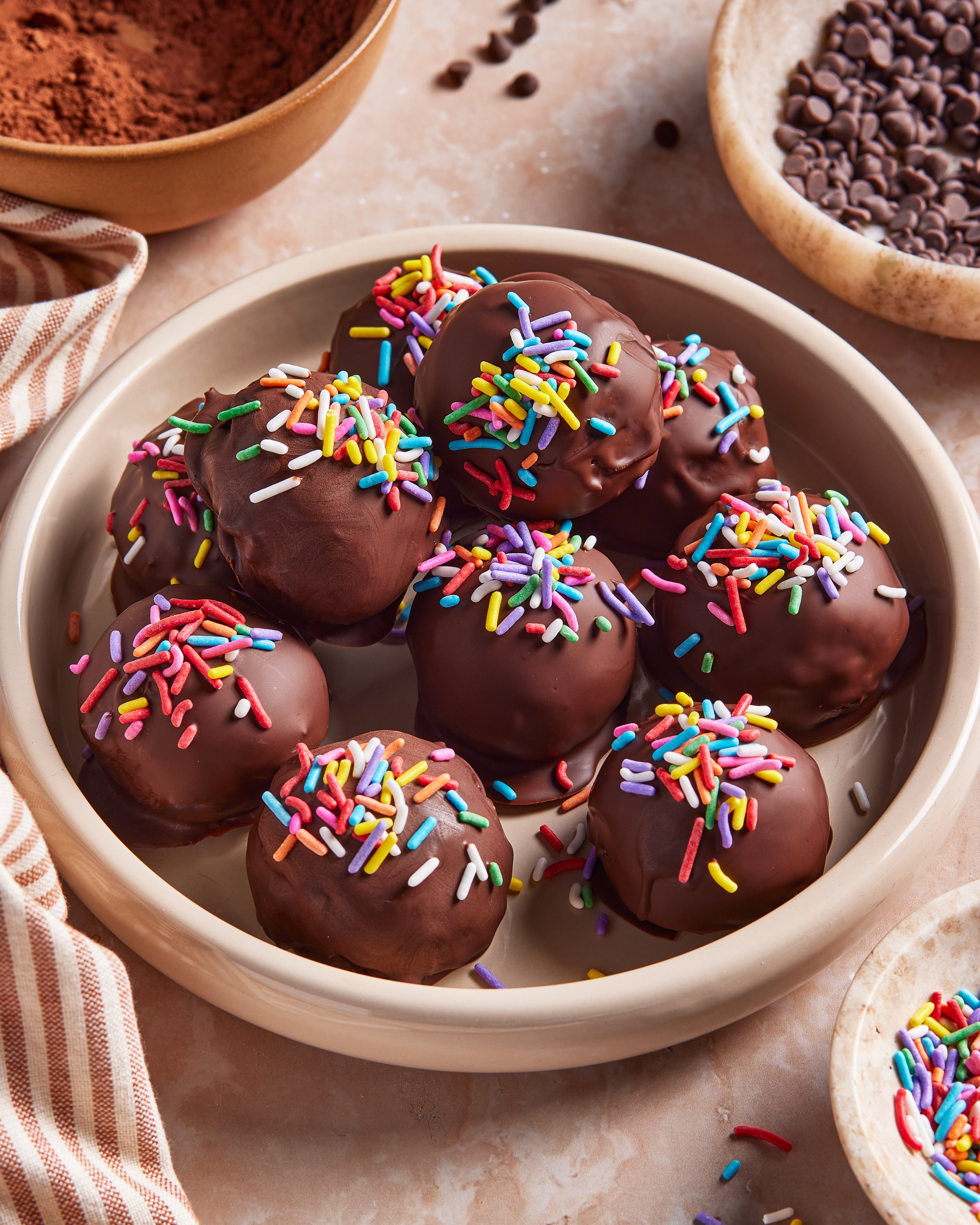 A bowl filled with chocolate-coated balls topped with colorful sprinkles sits on a marble surface, surrounded by bowls of chocolate chips, cocoa powder, and a striped cloth. (Viral High Protein Brownie Bites)