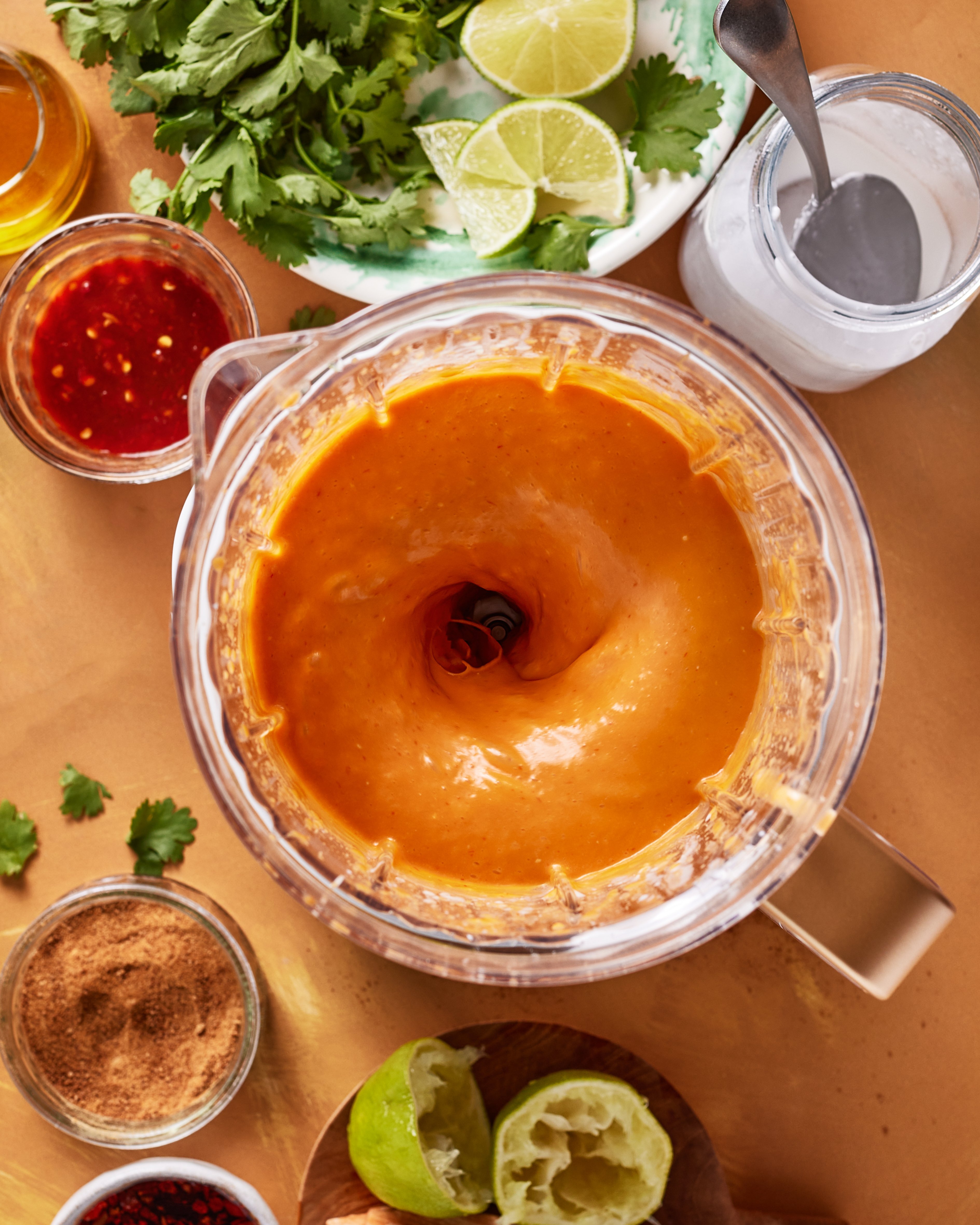 A blender filled with orange sauce sits on a table surrounded by fresh cilantro, lime wedges, crushed spices, chili sauce, coconut milk, and a squeezed lime half.