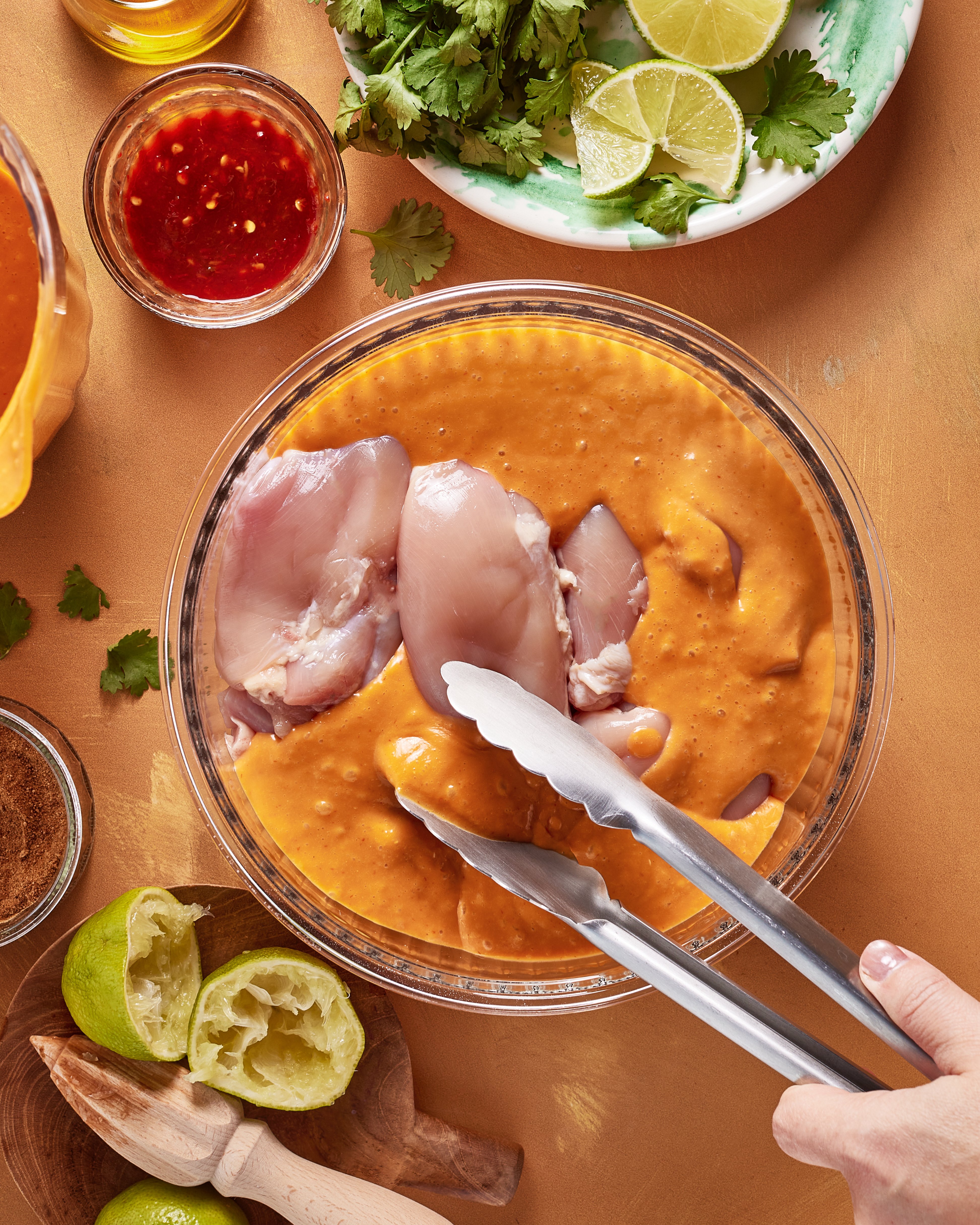 A hand uses tongs to dip raw chicken pieces into an orange marinade in a glass bowl. Surrounding items include squeezed limes, cilantro, lime wedges, chili sauce, and a spice bowl on a tan surface.
