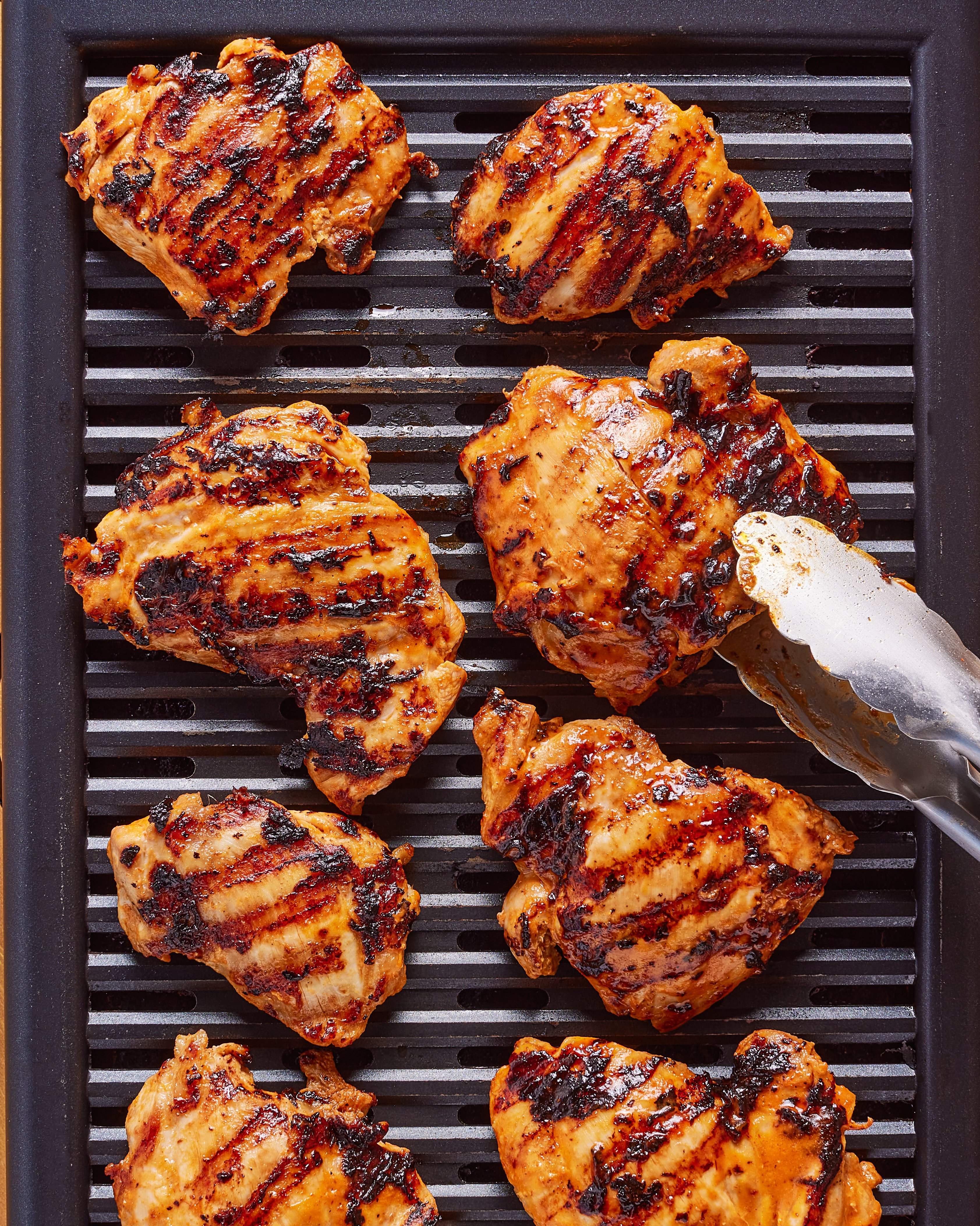 Grilled chicken thighs with visible char marks are arranged on a grill pan. Metal tongs are holding one piece, suggesting the chicken is being turned or removed from the grill.