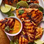 Grilled chicken thighs served on a white platter with lime wedges, fresh cilantro, and a small bowl of orange dipping sauce. The chicken has visible grill marks and a golden, slightly charred appearance.