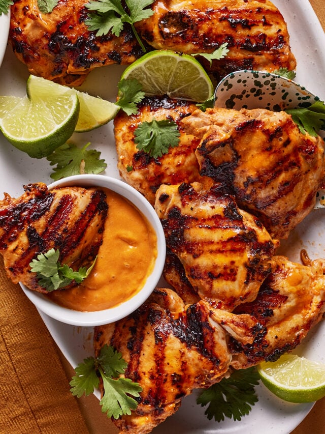 Grilled chicken thighs served on a white platter with lime wedges, fresh cilantro, and a small bowl of orange dipping sauce. The chicken has visible grill marks and a golden, slightly charred appearance.