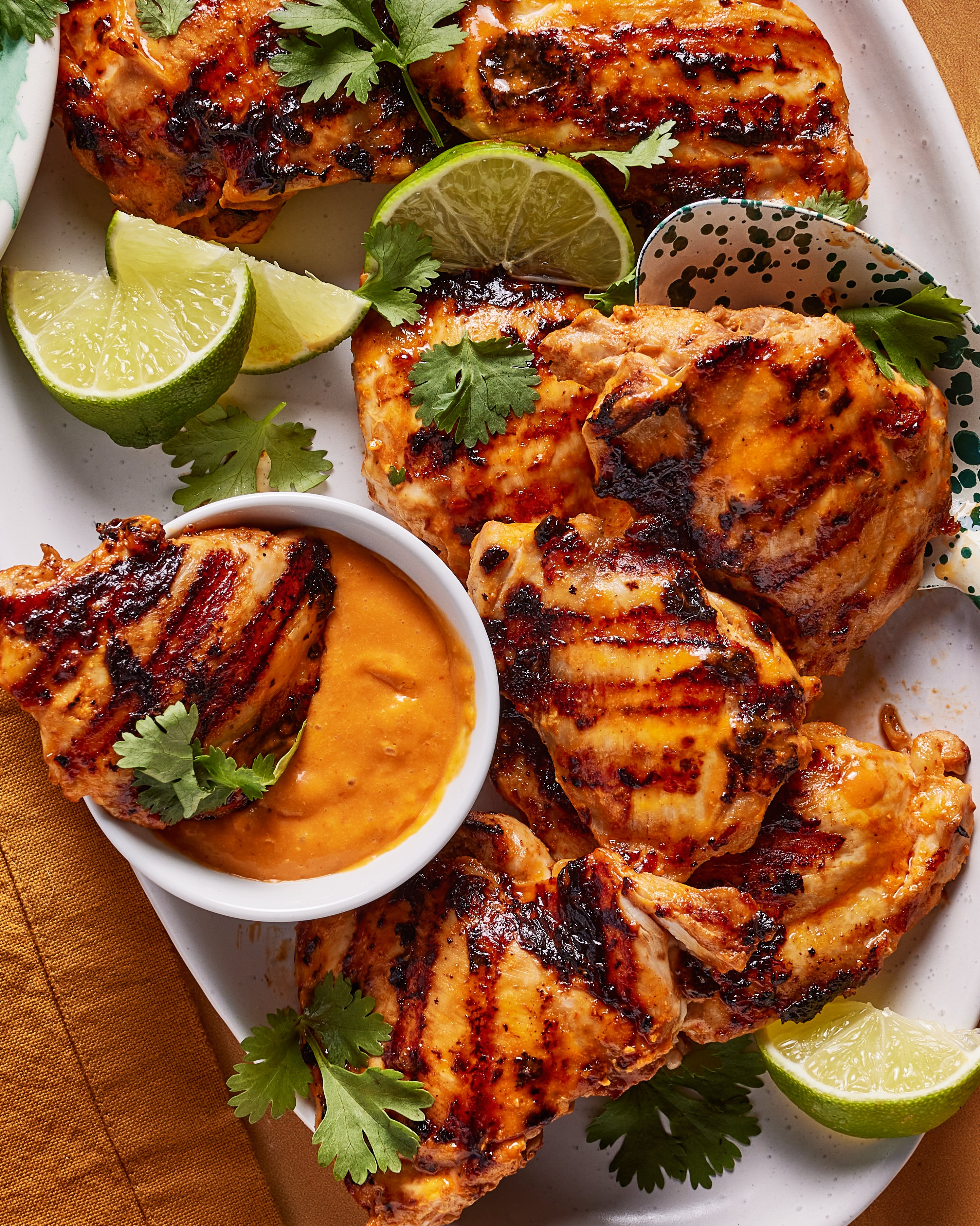 Grilled chicken thighs served on a white platter with lime wedges, fresh cilantro, and a small bowl of orange dipping sauce. The chicken has visible grill marks and a golden, slightly charred appearance.