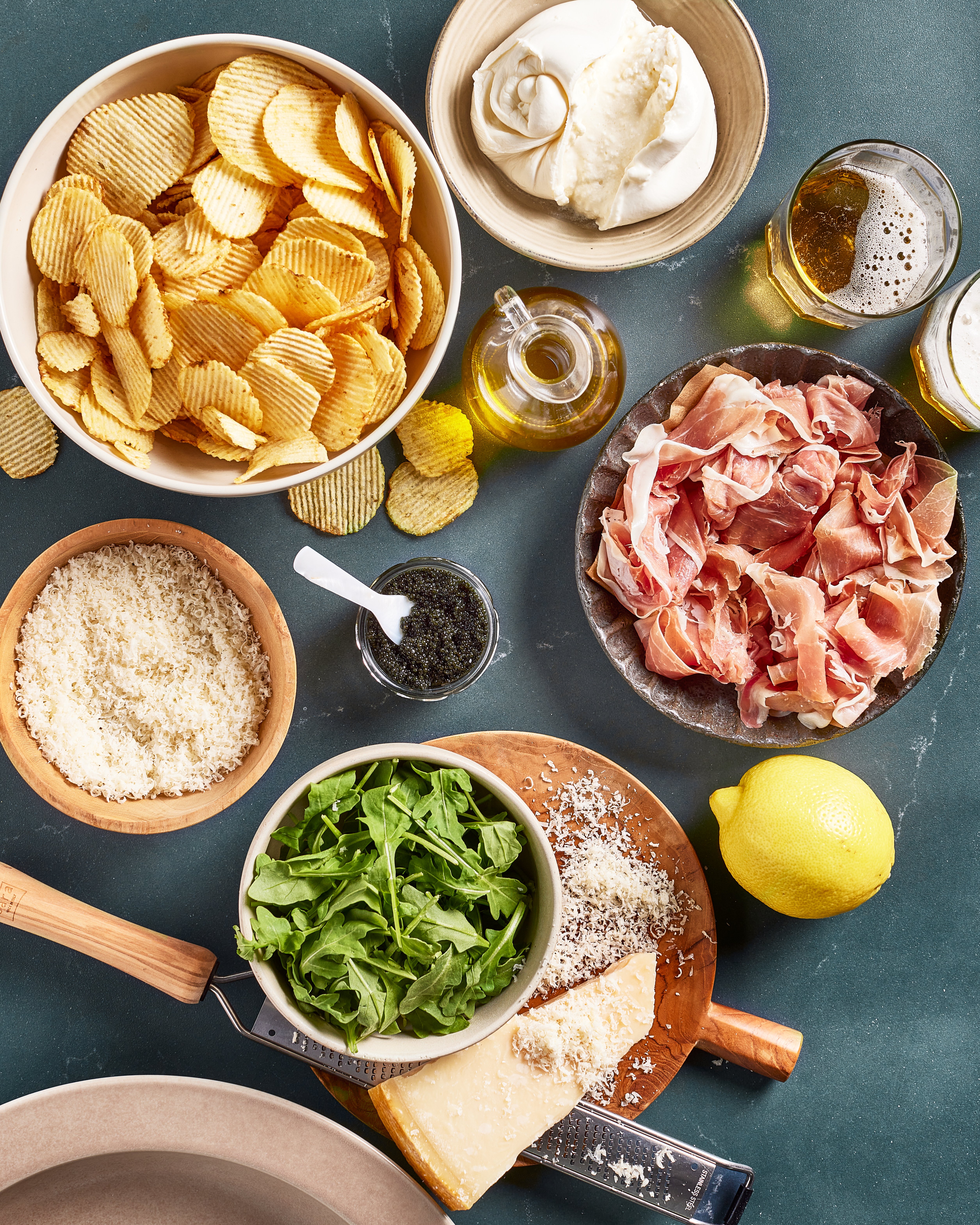 An overhead view of snacks and ingredients: a bowl of ridged potato chips, prosciutto, parmigiano cheese with a grater, arugula, a lemon, a dish of pesto, olive oil, a bowl of creamy dip, and glasses of beer on a green surface.