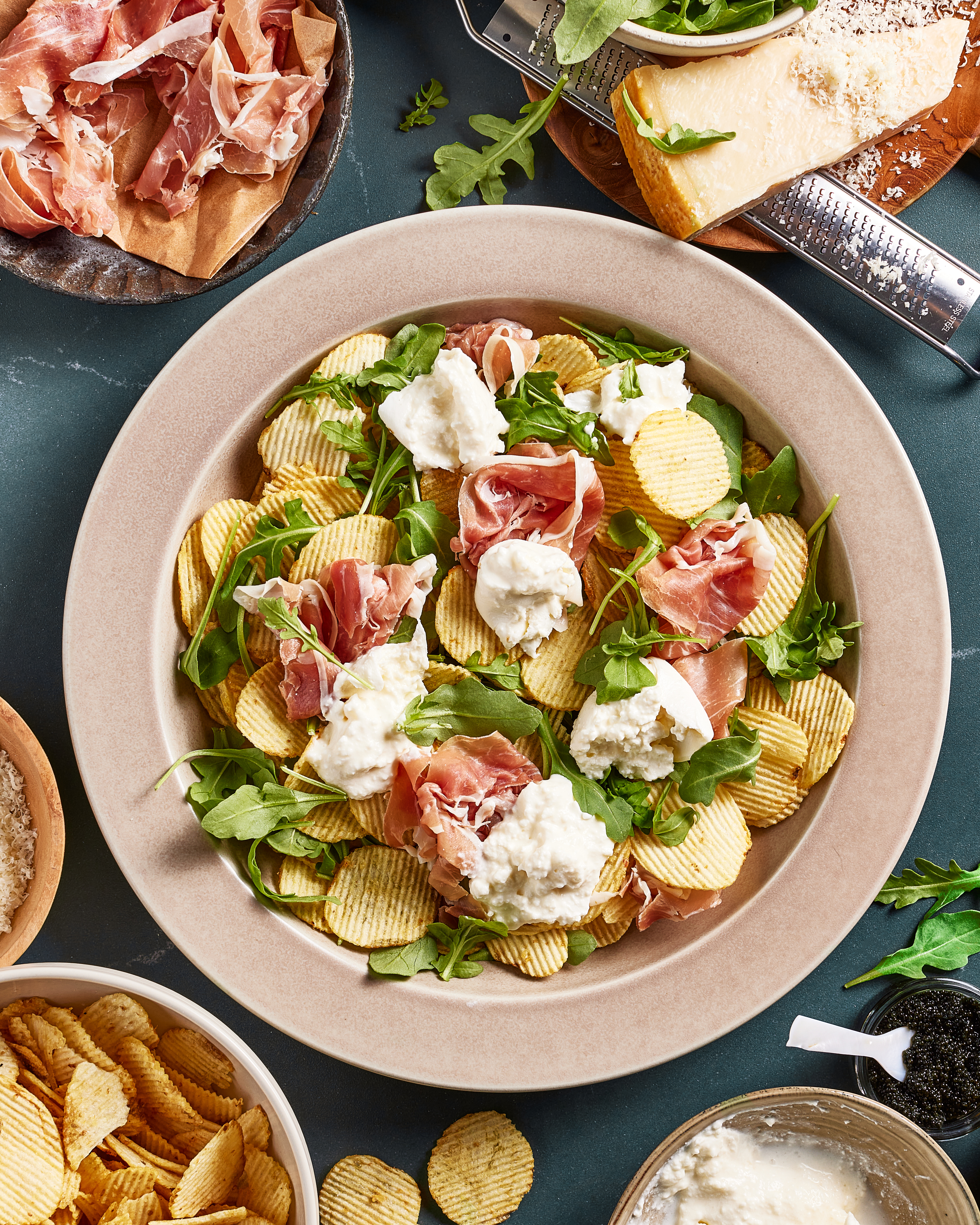 A plate of ridged potato chips topped with arugula, prosciutto, and dollops of burrata cheese, surrounded by bowls of chips, parmesan cheese, and a cheese grater on a dark surface.