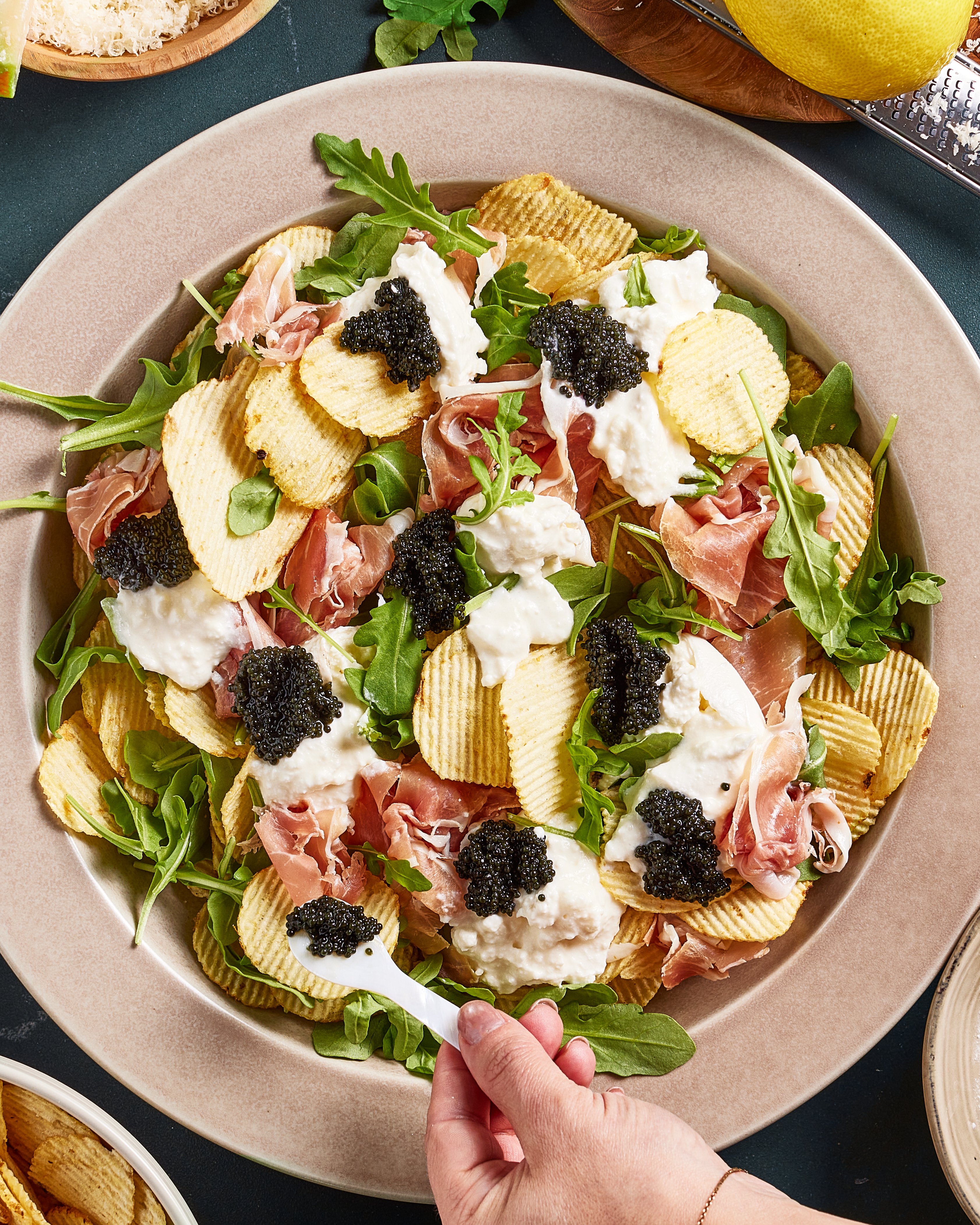 A hand adds black caviar to a salad with arugula, ridged potato chips, burrata cheese, and prosciutto, served on a beige plate.