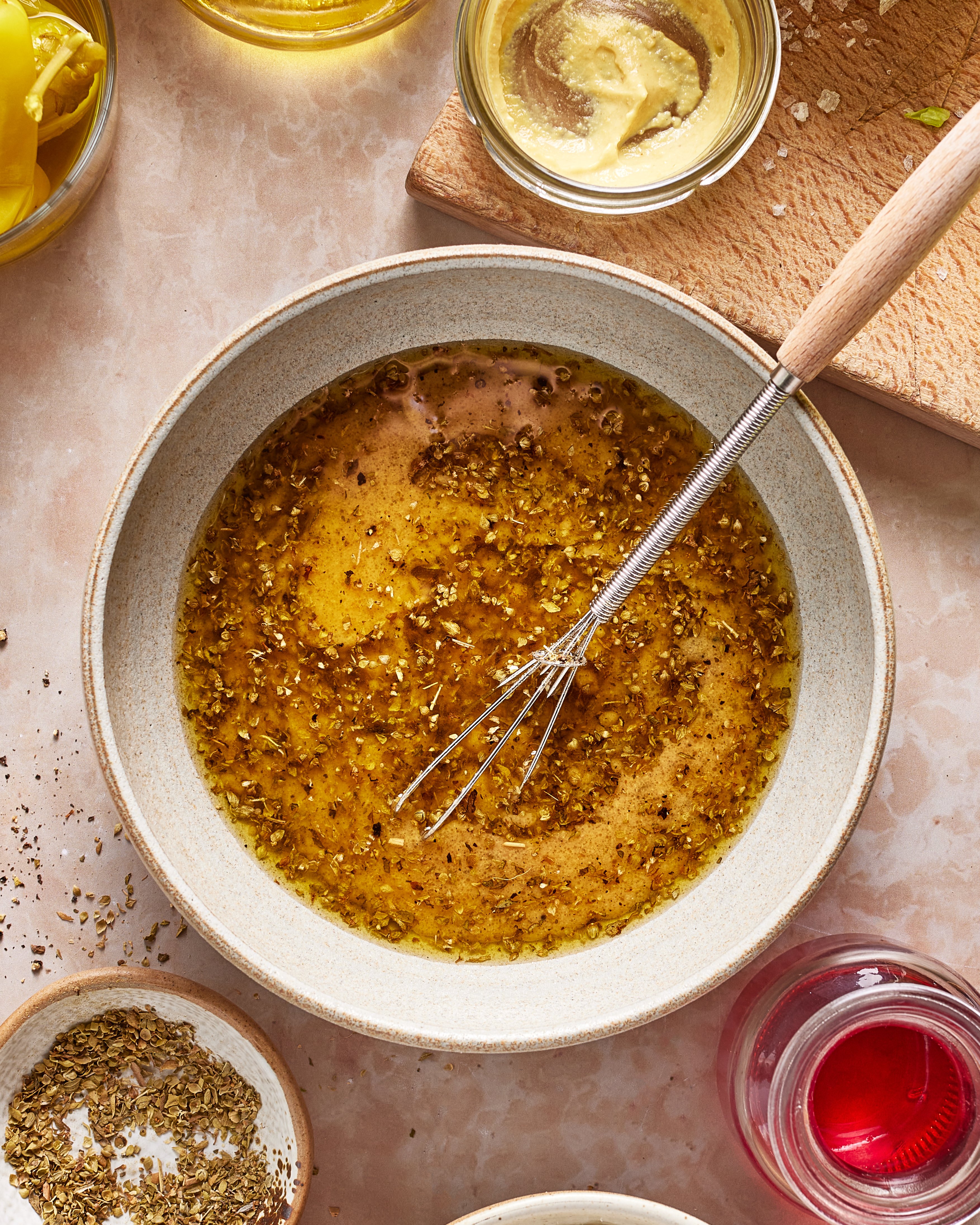 A bowl of mixed vinaigrette with herbs is whisked on a counter, surrounded by ingredients including mustard, oil, and vinegar, plus a cutting board.