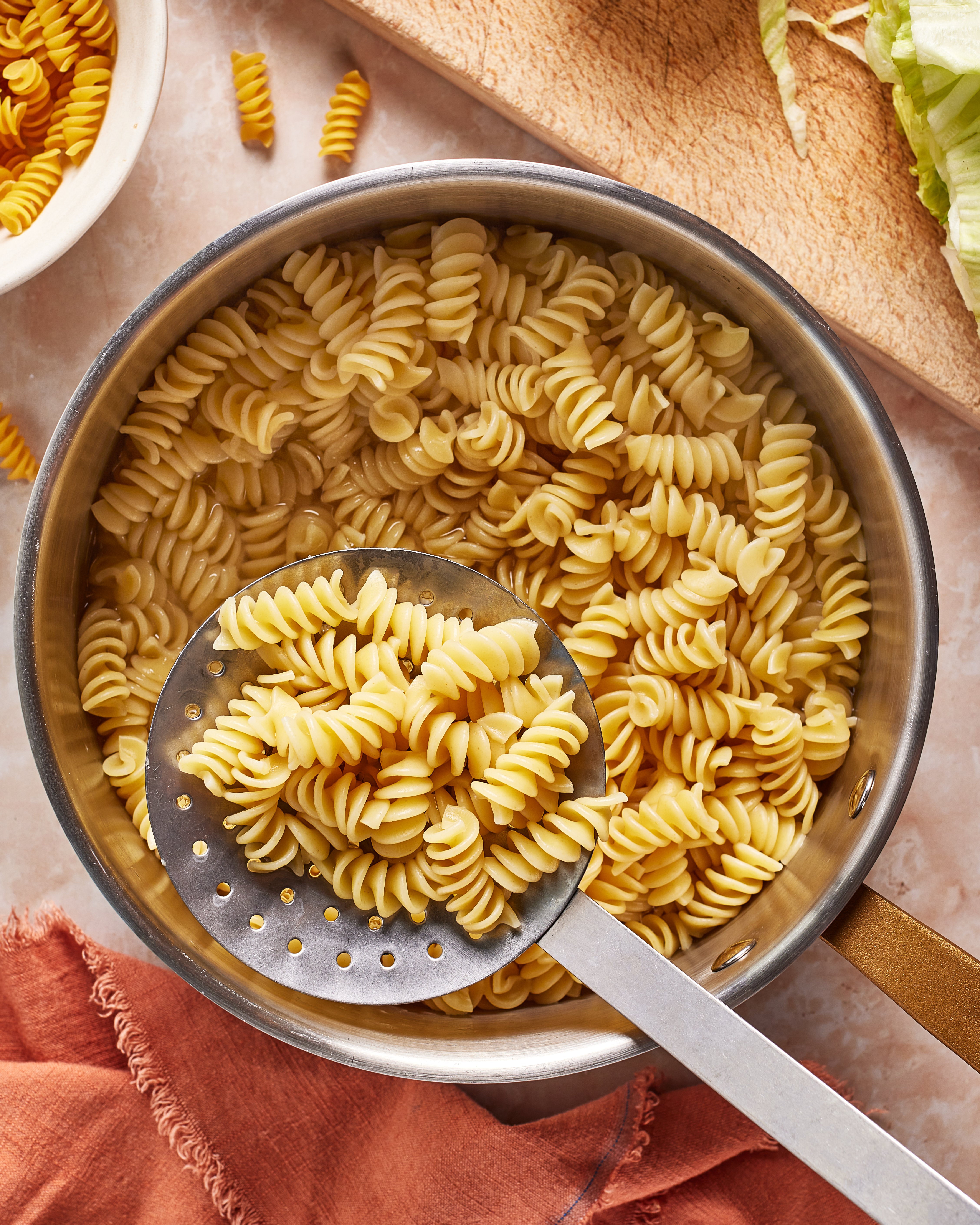 A saucepan filled with cooked rotini pasta is shown from above. A metal slotted spoon rests in the pan, lifting some of the pasta. A wooden cutting board and a rust-colored cloth are nearby.