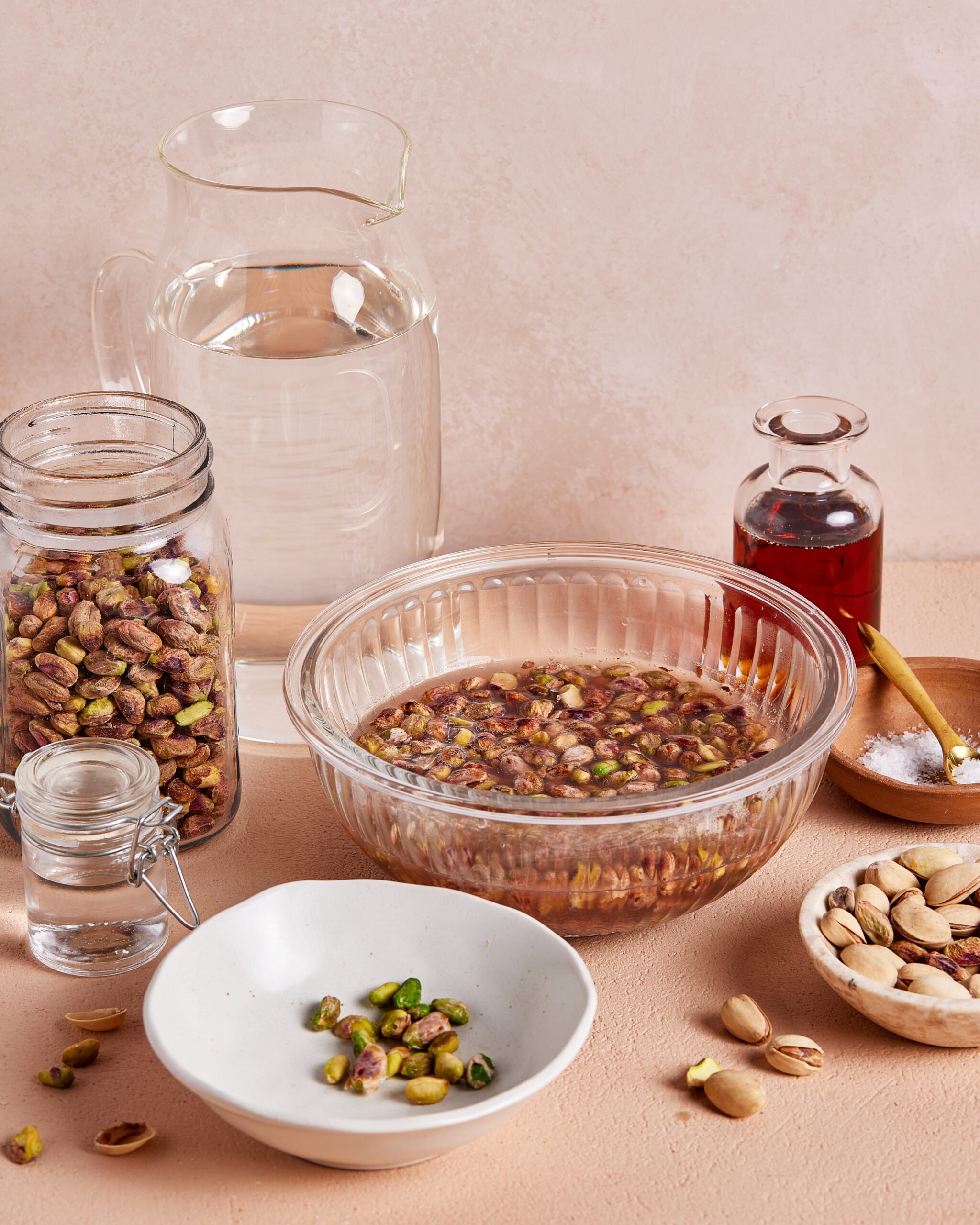 A glass bowl of pistachios soaking in water is surrounded by a jar of pistachios, a jug of water, a small bottle of rose water, a bowl of shelled pistachios, salt, and a dish with chopped pistachios.