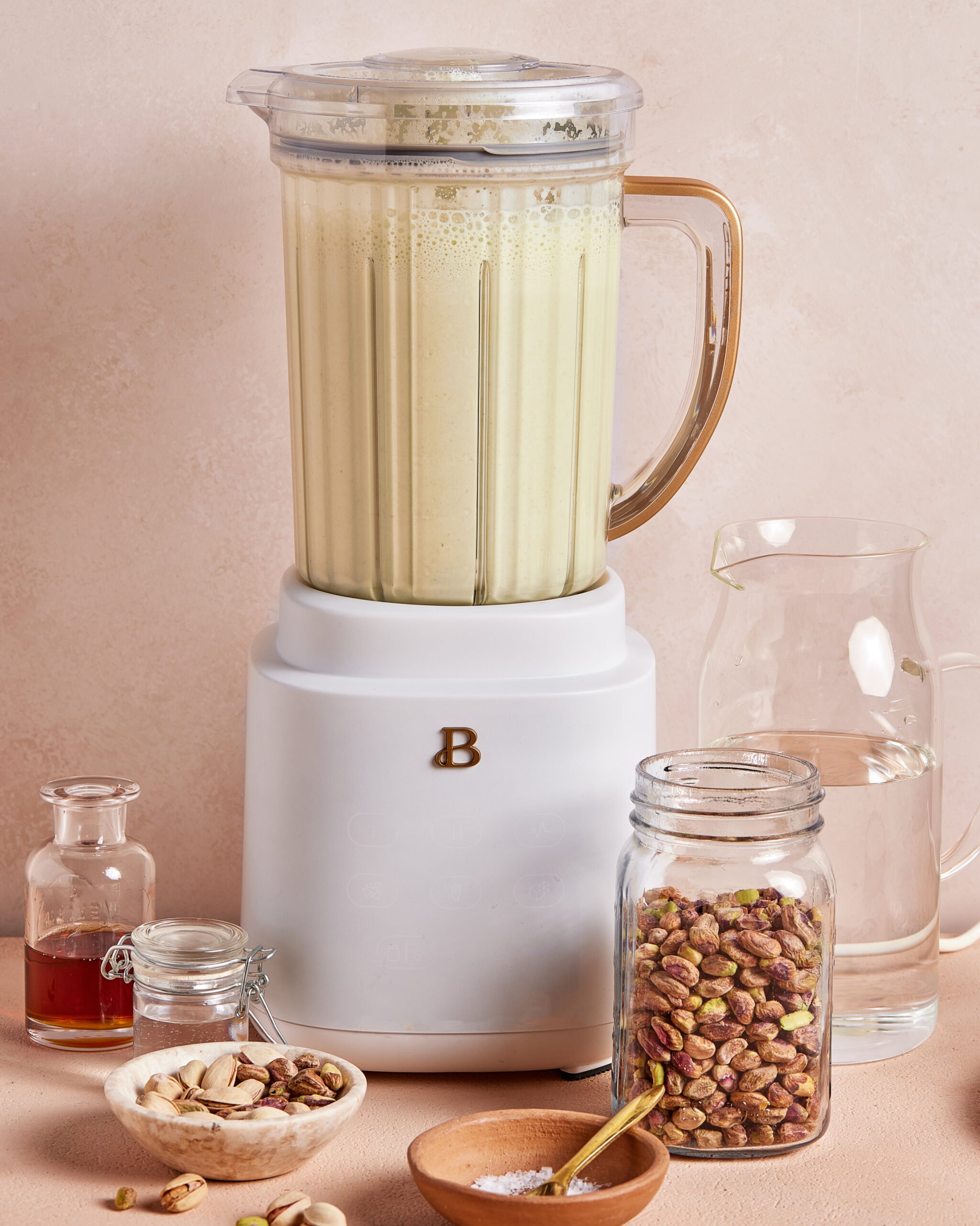 A blender filled with a pale green smoothie sits on a counter, surrounded by glass jars of pistachios, honey, salt, and a pitcher of water. Some pistachios are scattered on the surface.