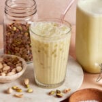 A glass of iced pistachio milk with a glass straw sits on a marble surface, surrounded by a jar of pistachios, a bowl of salt, scattered pistachios, and a bottle of pistachio milk.