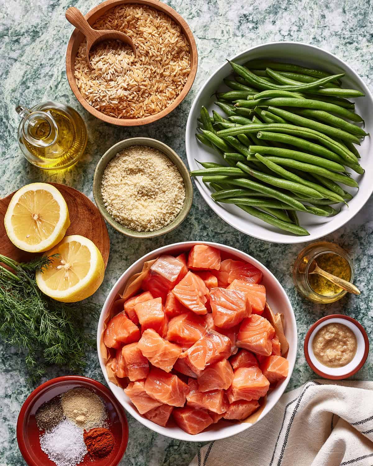 A top-down view of bowls containing cubed salmon, green beans, brown rice, grated Parmesan, olive oil, sliced lemons, minced garlic, fresh dill, and assorted spices on a green marbled surface.