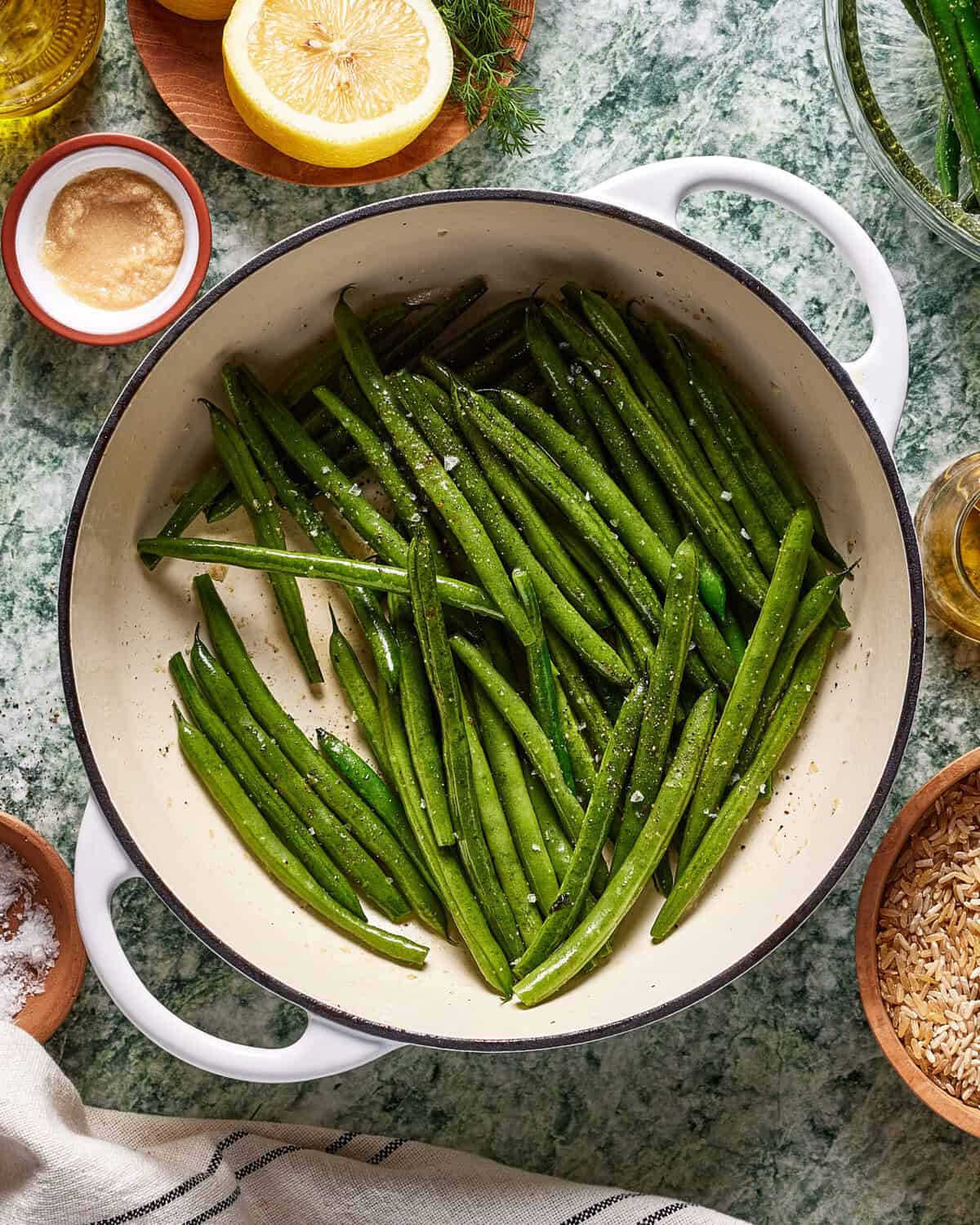 A white pot filled with fresh green beans sprinkled with salt, surrounded by bowls of ingredients, a lemon half, and a striped towel on a green marble surface.
