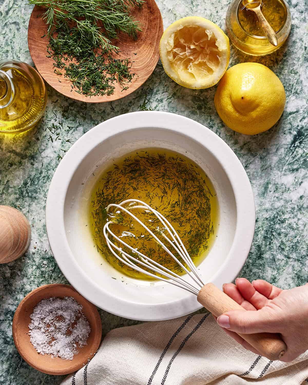 A hand whisks olive oil, chopped herbs, and lemon juice in a white bowl on a green countertop, surrounded by fresh dill, lemons, salt, a towel, and bottles of olive oil.