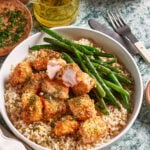 A bowl of brown rice topped with crispy breaded crispy salmon bites, fresh green beans, and sprinkled herbs. A fork and knife are beside the bowl, with a small jar of olive oil and a dish of herbs in the background.