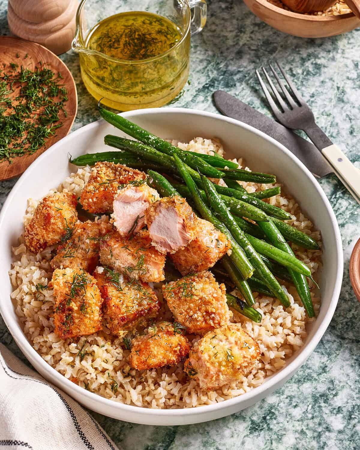A bowl of brown rice topped with crispy breaded crispy salmon bites, fresh green beans, and sprinkled herbs. A fork and knife are beside the bowl, with a small jar of olive oil and a dish of herbs in the background.