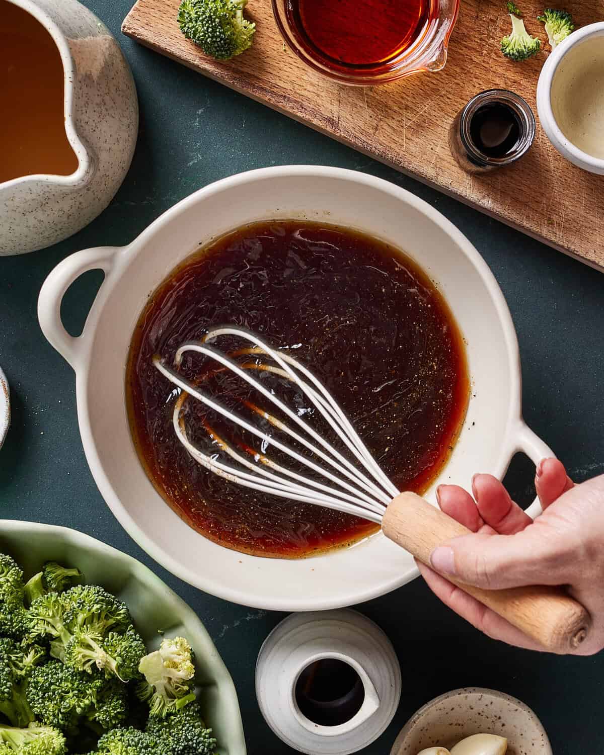 A hand whisking a dark sauce in a white bowl, surrounded by bowls of broccoli, sauces, and seasonings on a green countertop.
