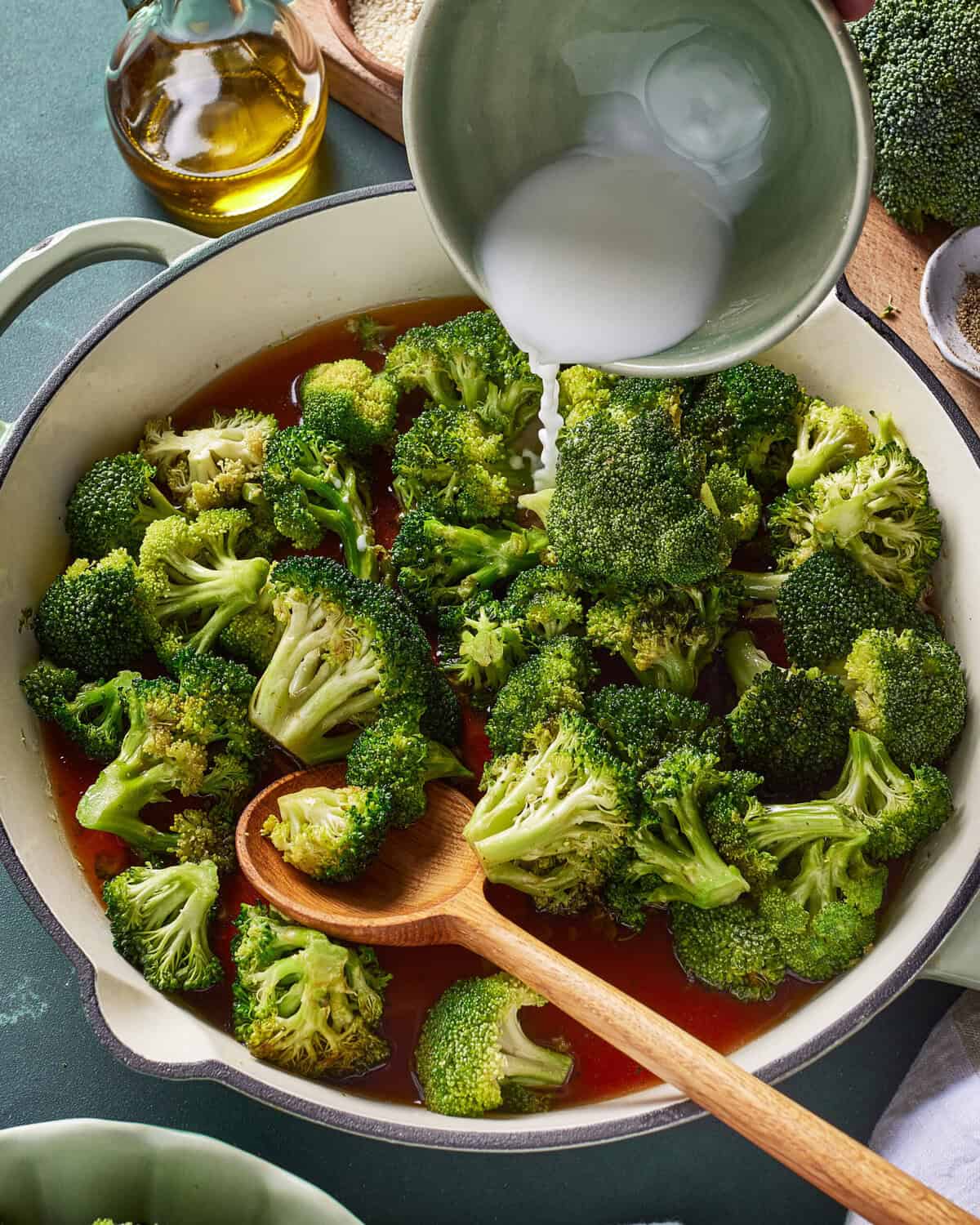 A pot filled with steamed broccoli and sauce is being stirred with a wooden spoon, while a person pours a white liquid, likely a sauce or broth, from a bowl into the pot. Cooking ingredients are visible in the background.