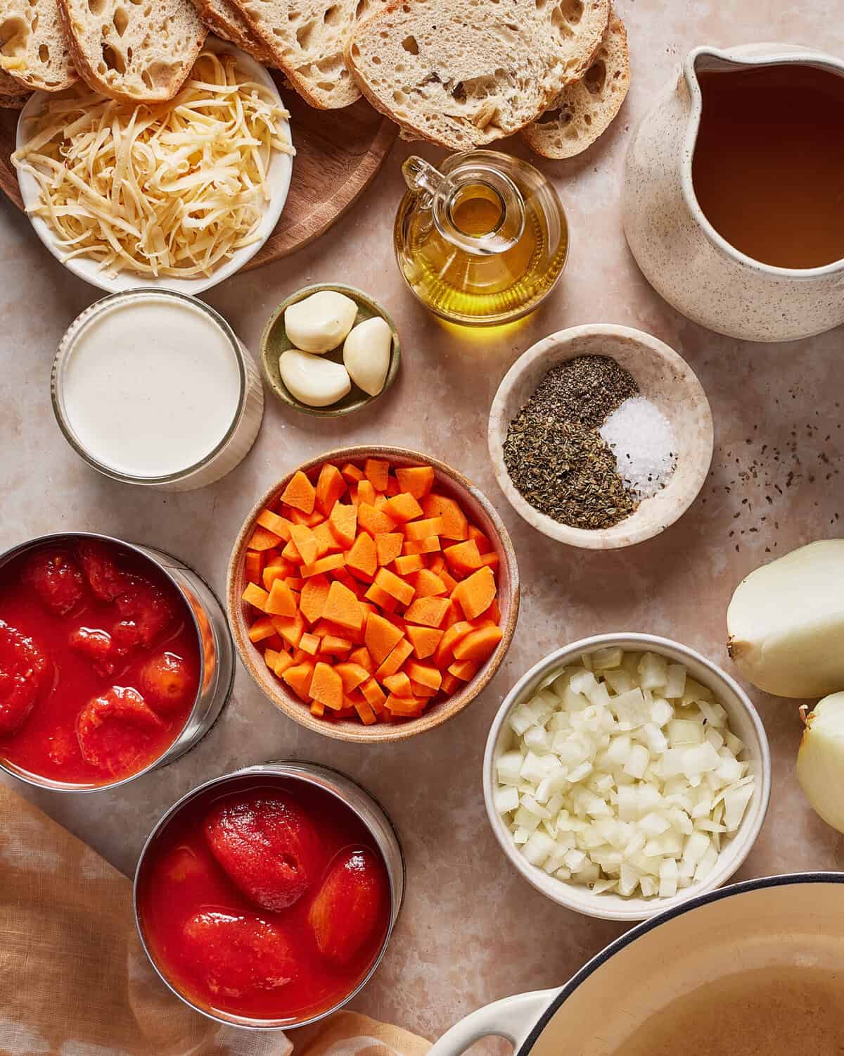 A flat lay of ingredients including chopped carrots, diced onions, garlic cloves, shredded cheese, canned tomatoes, olive oil, cream, dried herbs, salt, sliced bread, and a jug of broth on a light surface.