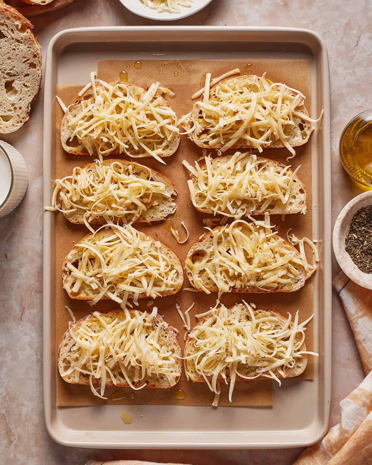 Nine slices of bread topped with shredded cheese and herbs are arranged on a parchment-lined baking tray, ready to be baked. Cooking ingredients and cloth napkins are visible around the tray.