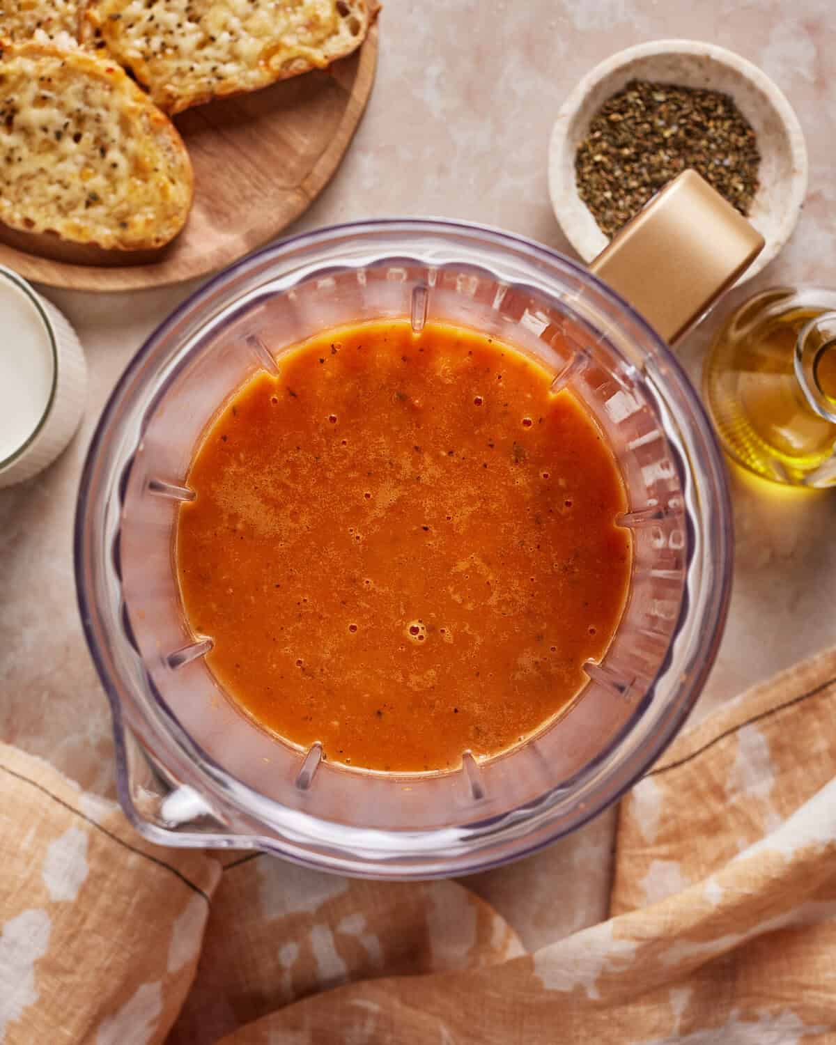 A blender filled with creamy tomato soup on a countertop, surrounded by a glass of milk, a bowl of dried herbs, a bottle of olive oil, toasted bread slices, and a tan-patterned cloth.