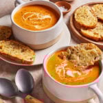 Two ceramic bowls of creamy tomato soup with a swirl of cream, served with slices of toasted bread topped with melted cheese. Spoons, pepper, and salt are placed nearby on a marble surface.