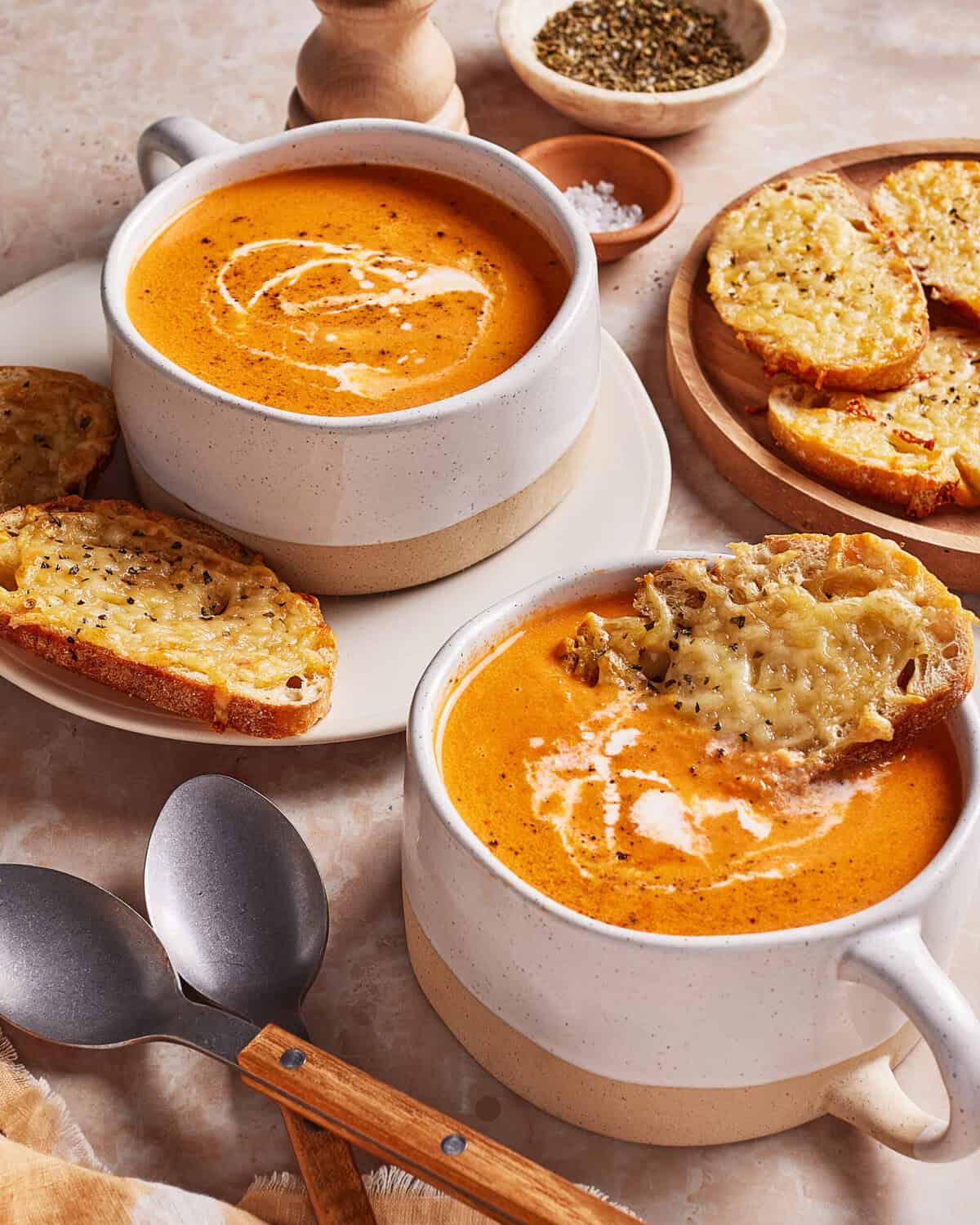 Two ceramic bowls of creamy tomato soup with a swirl of cream, served with slices of toasted bread topped with melted cheese. Spoons, pepper, and salt are placed nearby on a marble surface.