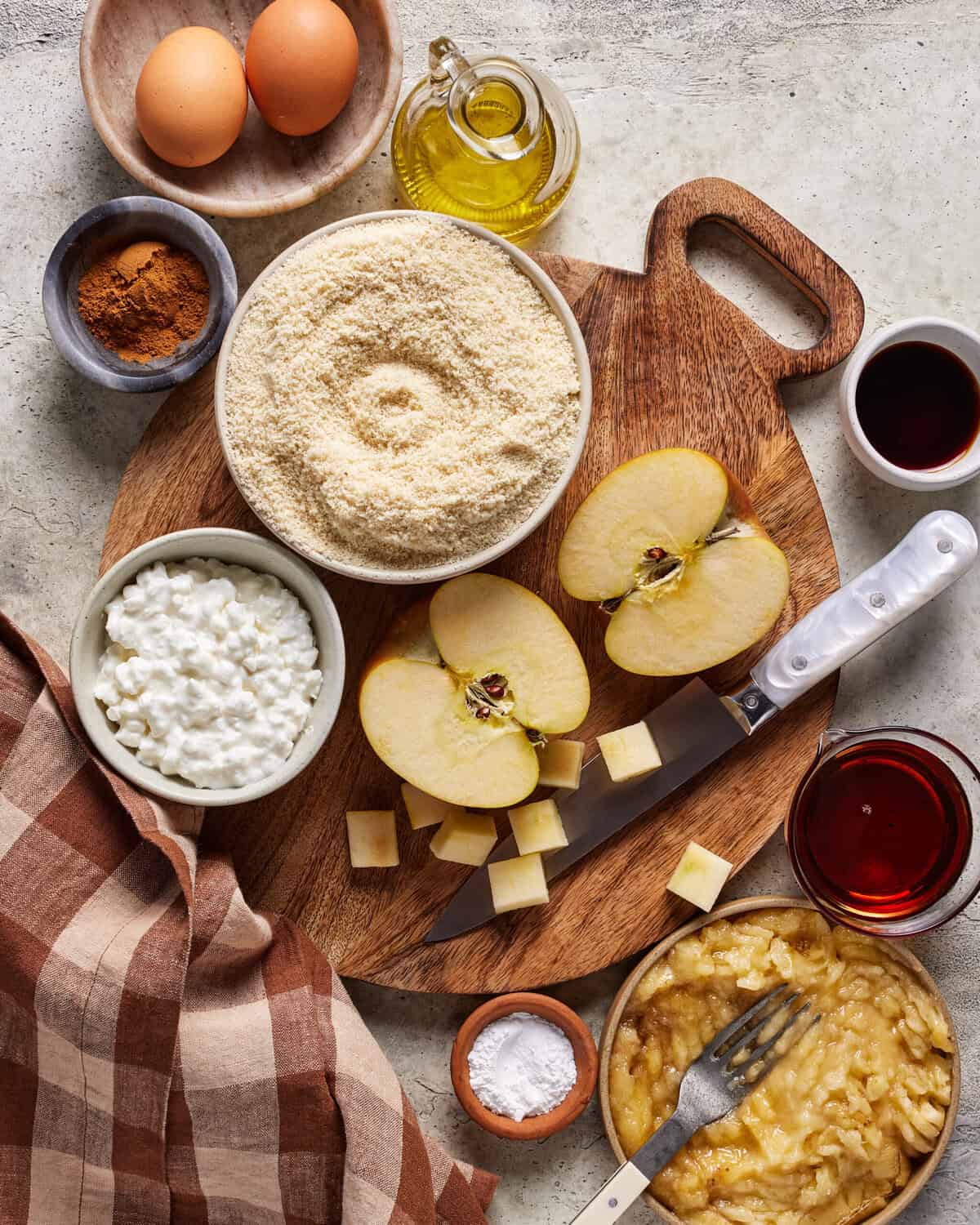 Ingredients for baking are arranged on a wooden board: eggs, flour, olive oil, cinnamon, cottage cheese, apple halves, apple cubes, vanilla, baking powder, applesauce, and a knife, with a checkered cloth on the side.