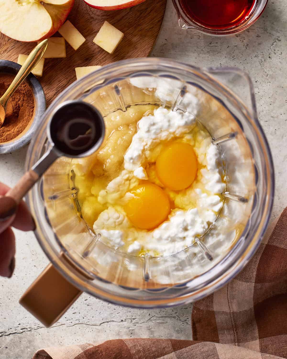 A blender contains two eggs, cottage cheese, and applesauce. A hand holds a tablespoon of vanilla over the blender. Nearby are apple slices, a bowl of cinnamon, and a cup of syrup on a light countertop.