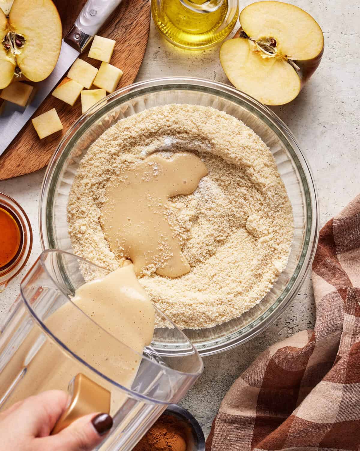 A hand pours a creamy mixture from a blender into a bowl of almond flour. Sliced apples, diced apple pieces, cinnamon, olive oil, and a brown cloth are on the table nearby.