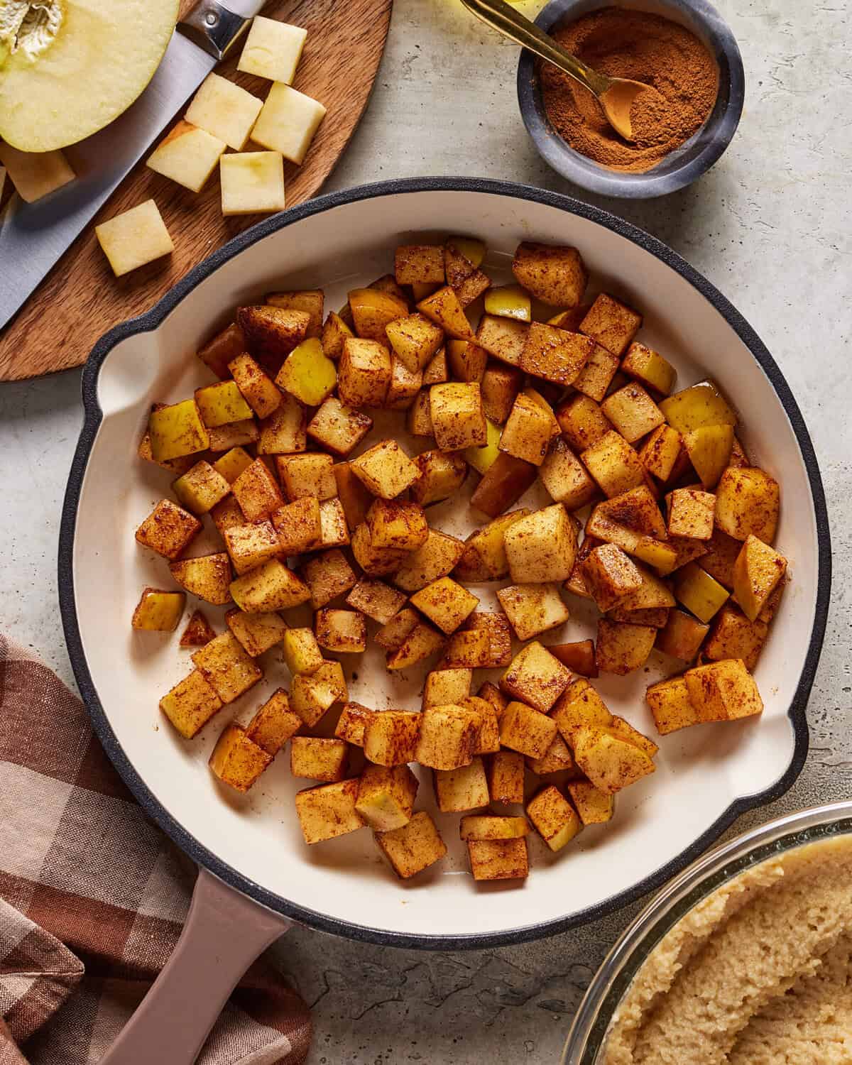 A skillet filled with diced apples coated in cinnamon sits on a countertop. Nearby are a bowl of cinnamon, a sliced apple, a knife, and a brown checkered kitchen towel.