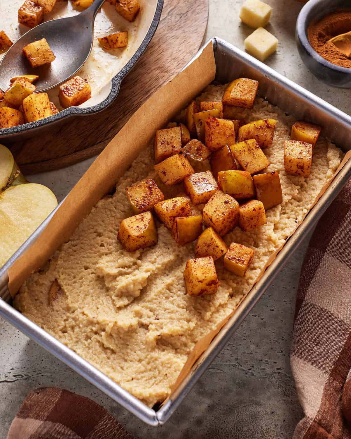 A baking pan with cake batter topped with spiced, cubed apples, ready to bake. Nearby are apple slices, a bowl of cinnamon, and a plaid cloth. A serving dish with more spiced apples is in the background.