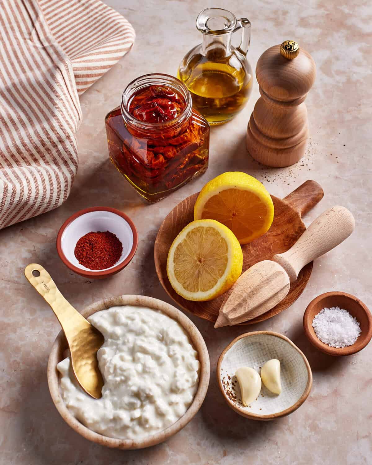 A bowl of cottage cheese with a gold spoon, lemon halves, a wooden citrus reamer, garlic cloves, a jar of sundried tomatoes in oil, a pepper grinder, salt, paprika, and a bottle of olive oil on a marble surface.