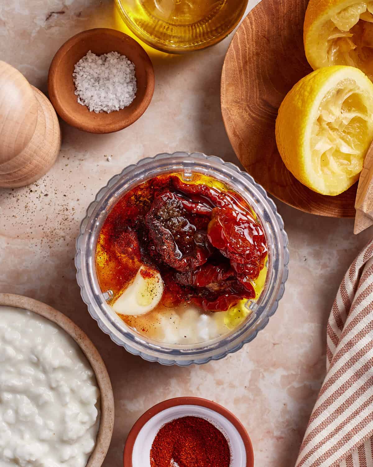 A glass bowl filled with yogurt, sun-dried tomatoes, garlic, paprika, and oil sits on a marble counter near cottage cheese, salt, paprika, a pepper mill, and squeezed lemons.